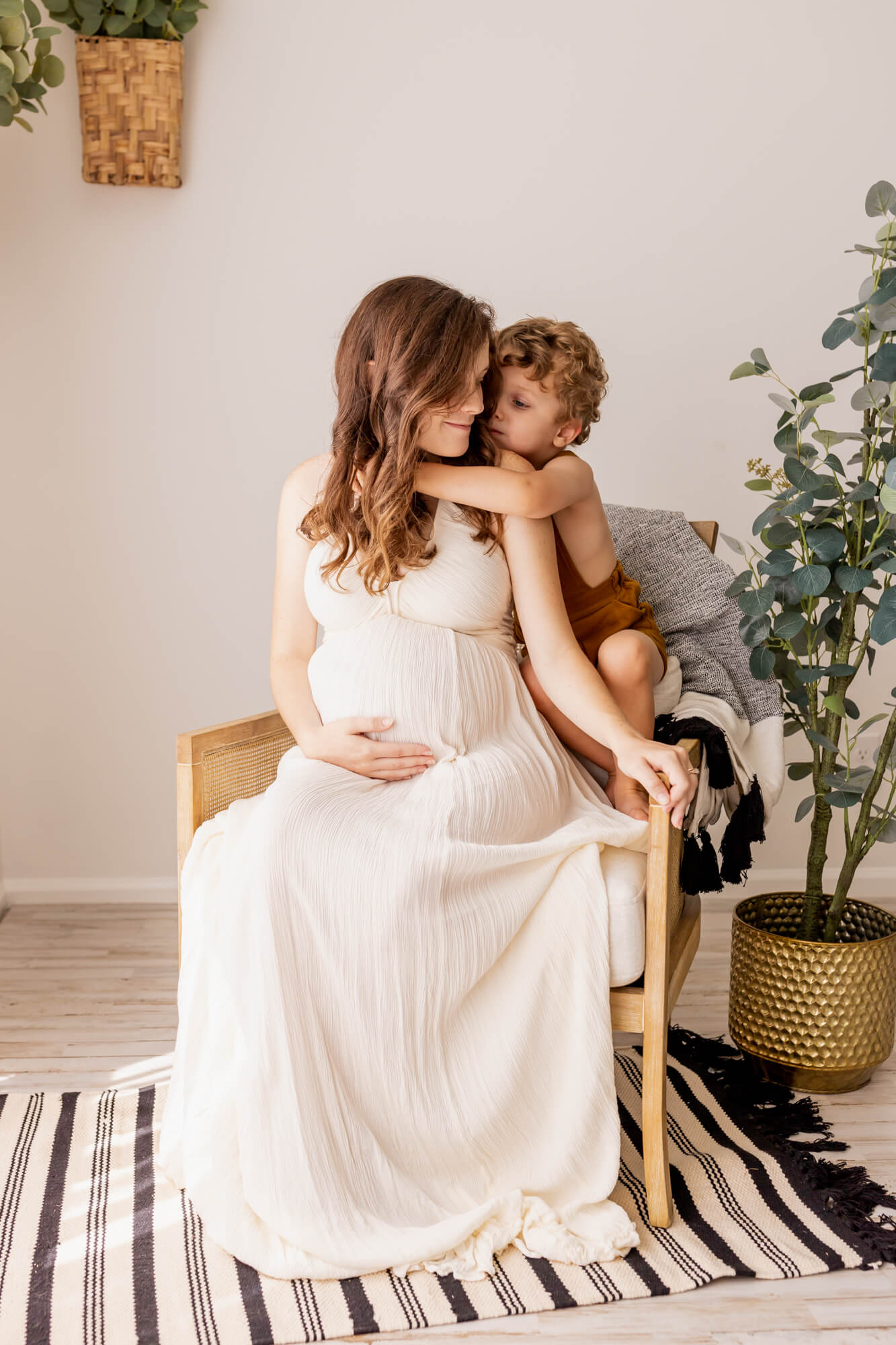pregnant woman in white gown sitting in a chair being hugged by her child