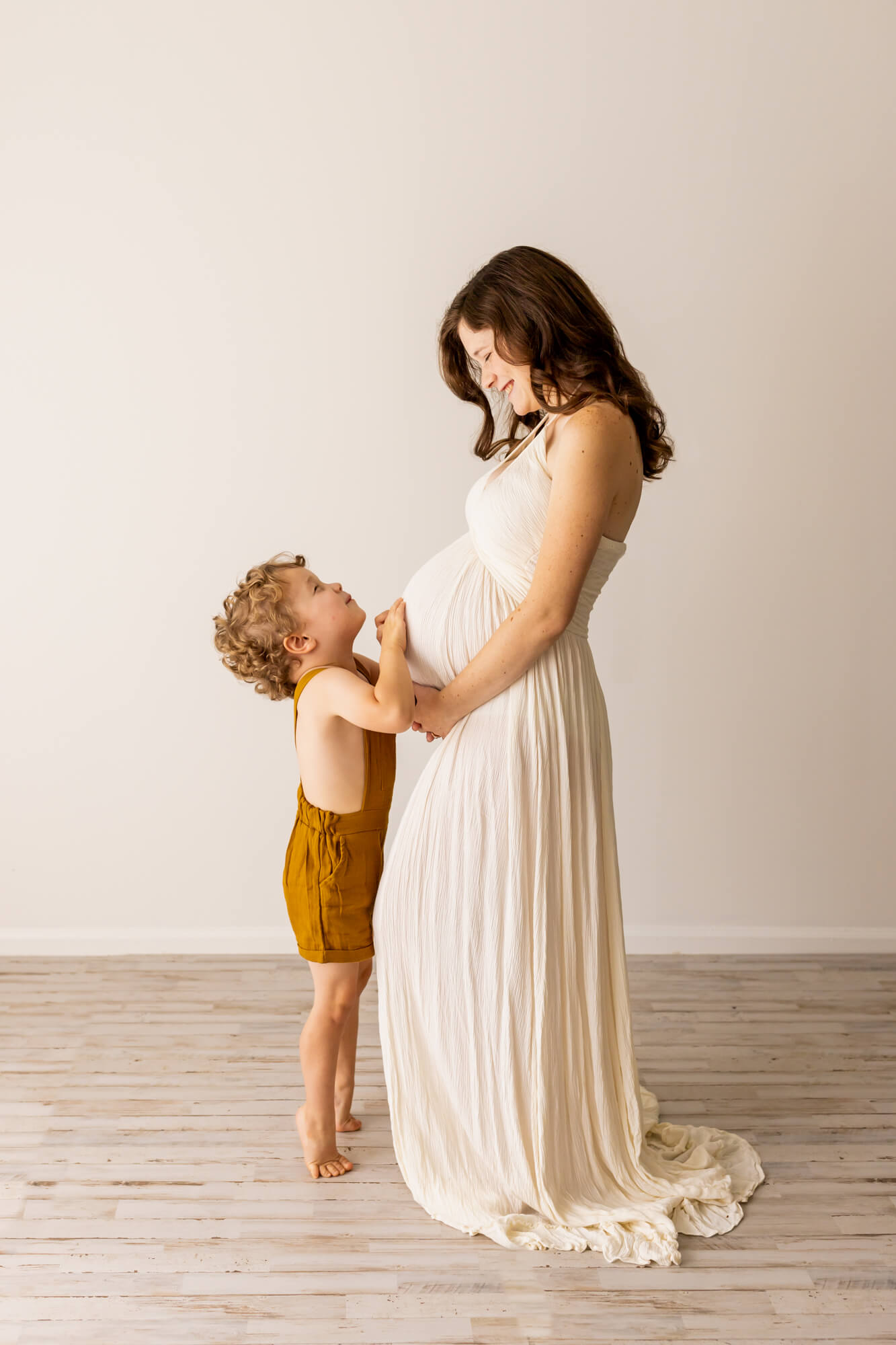 Mom to be in white gown holding her belly and looking at her son in brown overalls