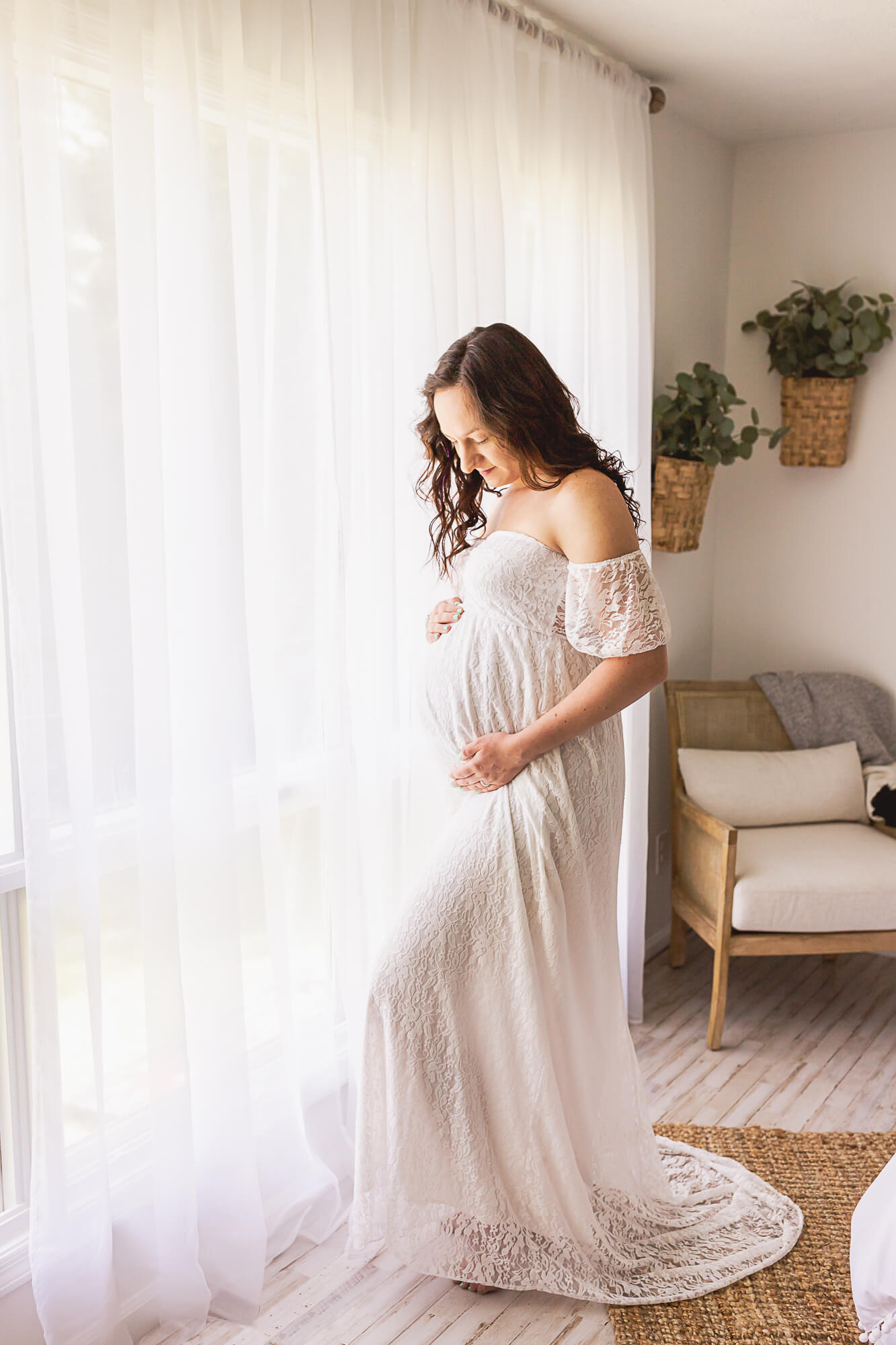 mom to be in off the shoulder white lace gown standing near a window