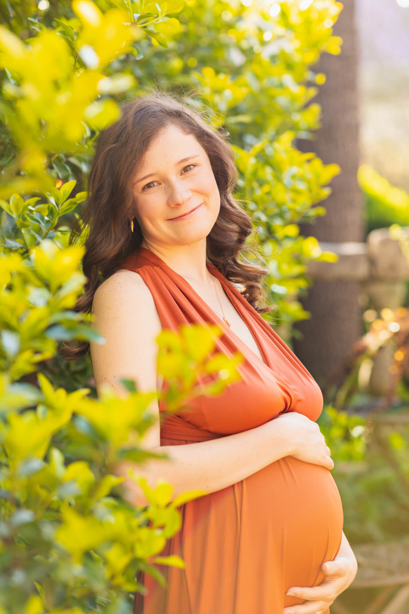 pregnant woman in orange maternity gown standing in greenery and smiling