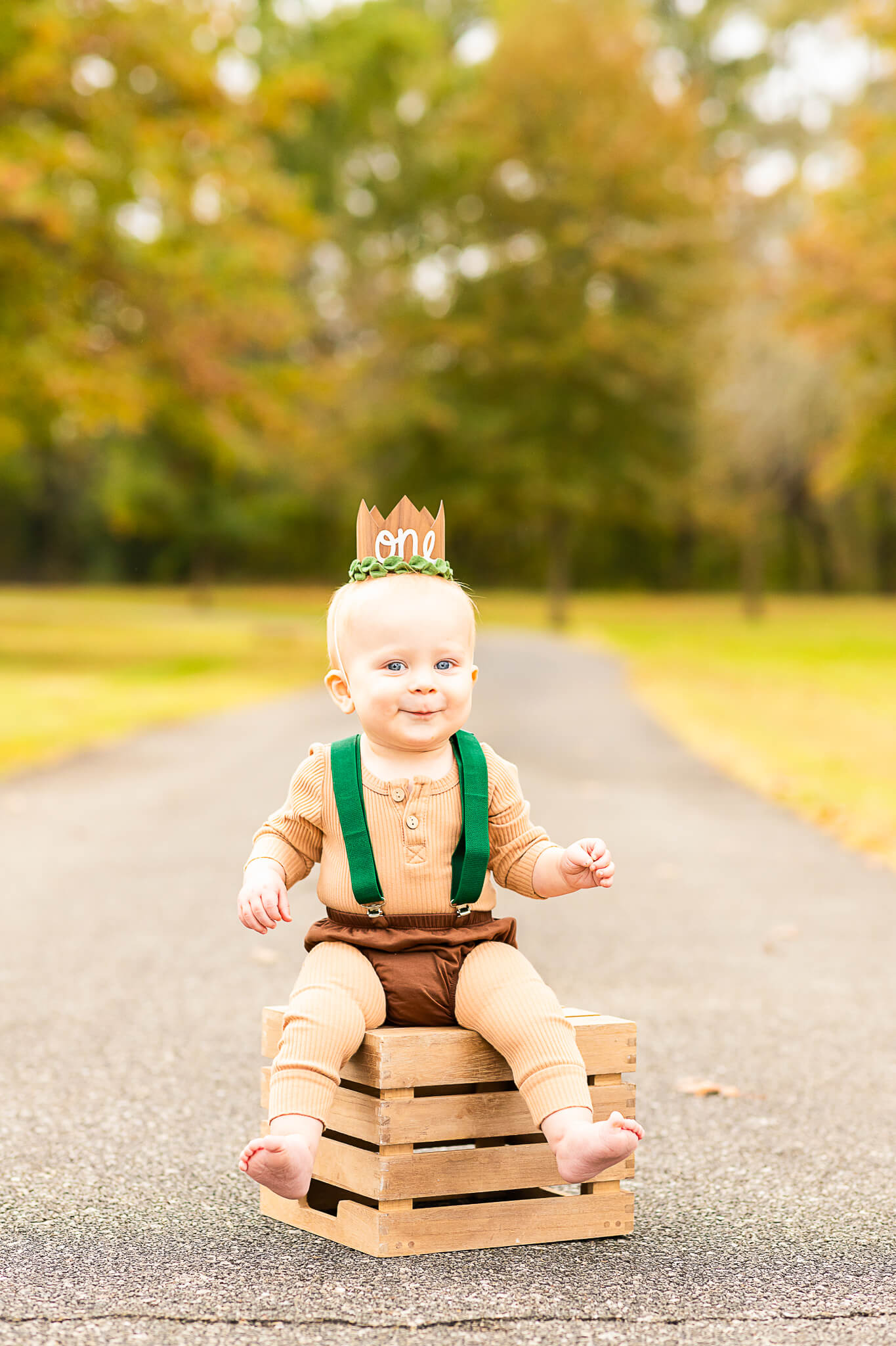 One year old boy sits on a wooden crate with a birthday hat in a park path