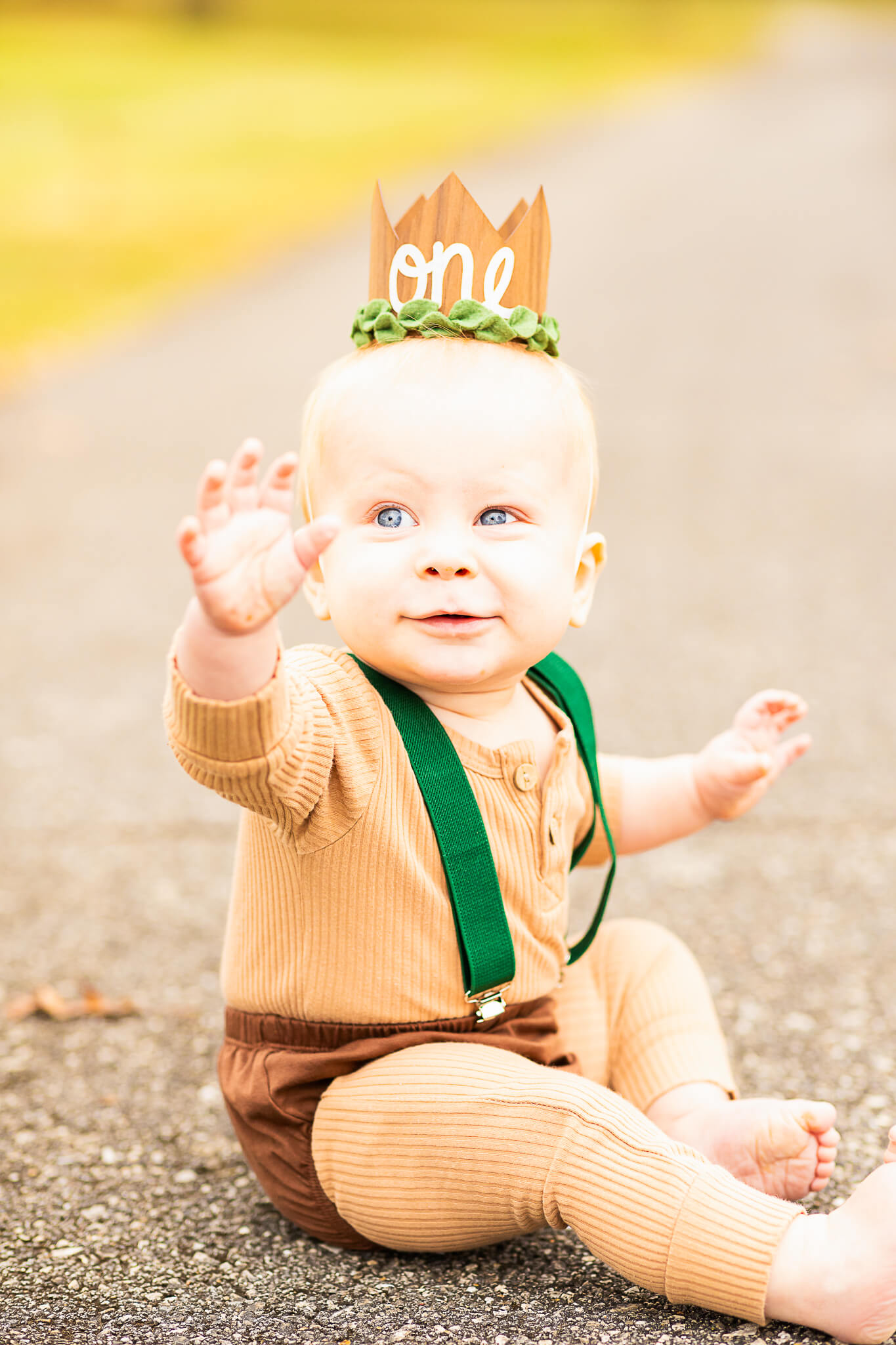 one year old boy sits in brown onesie and a birthday crown on a paved path birmingham cakes