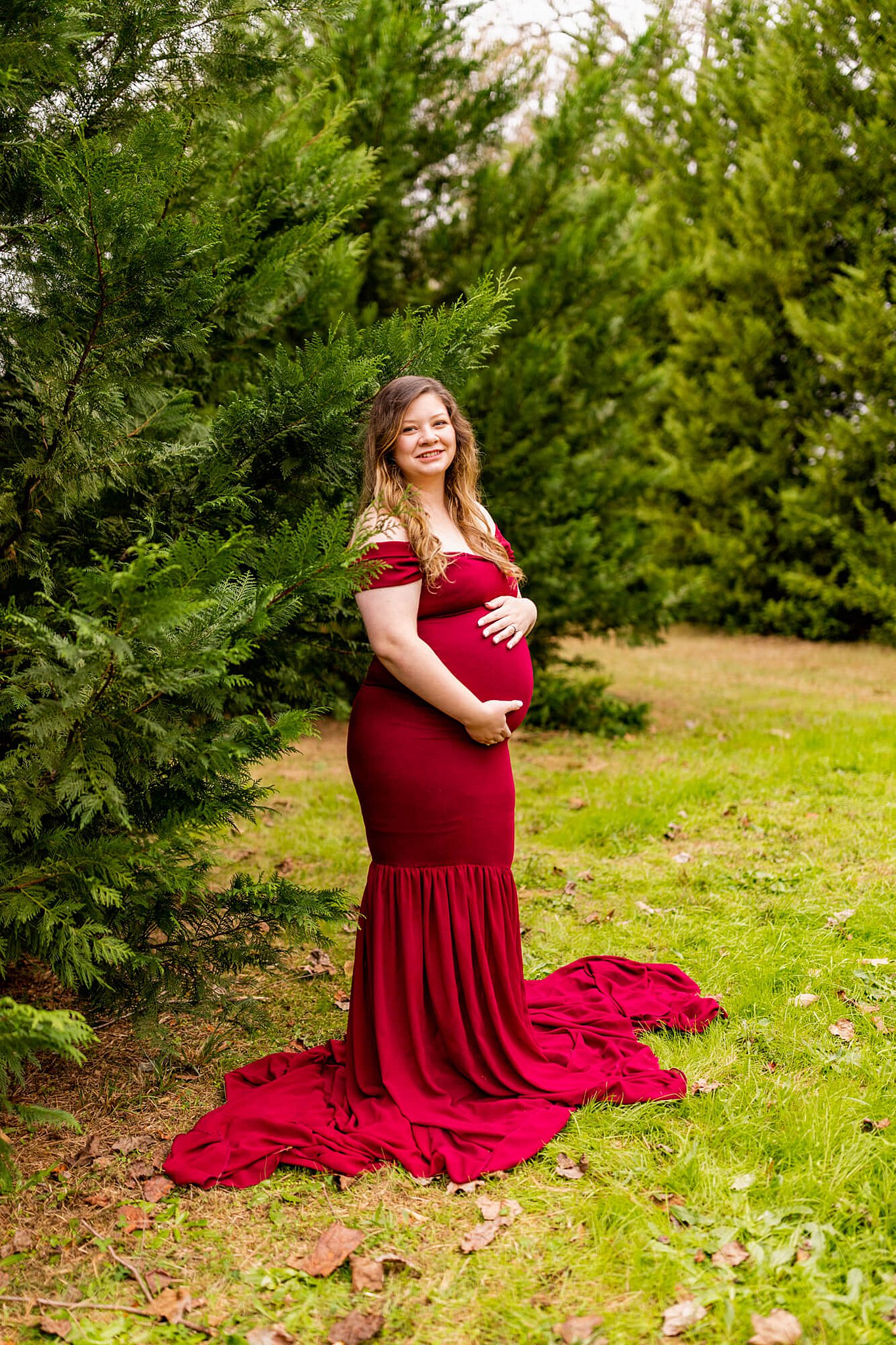 Mother-to-be stands by an evergreen tree holding her bump in a red maternity gown Brookwood Women’s Center