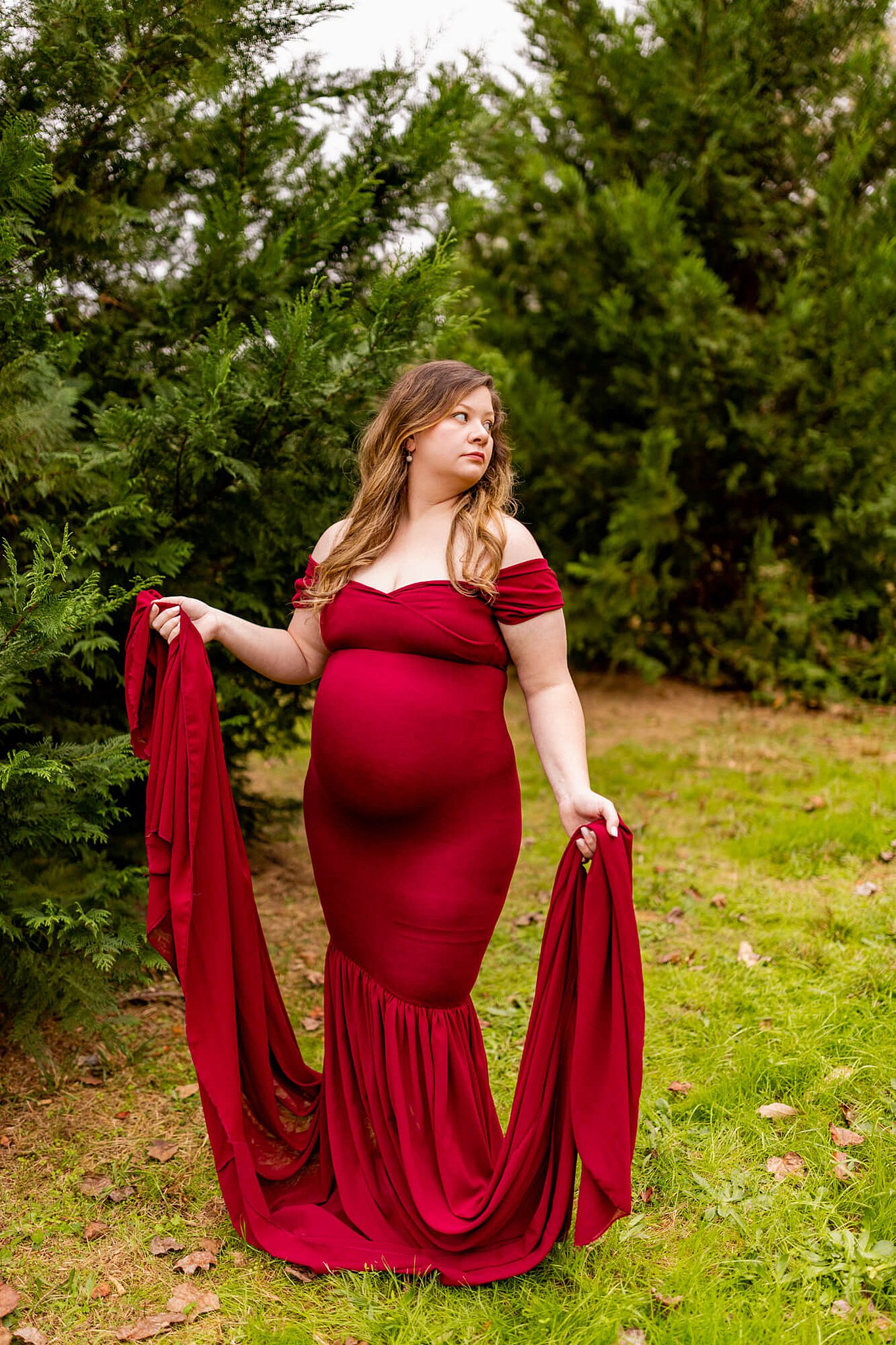 Mom-to-be holds the train of her maternity gown in an evergreen forest