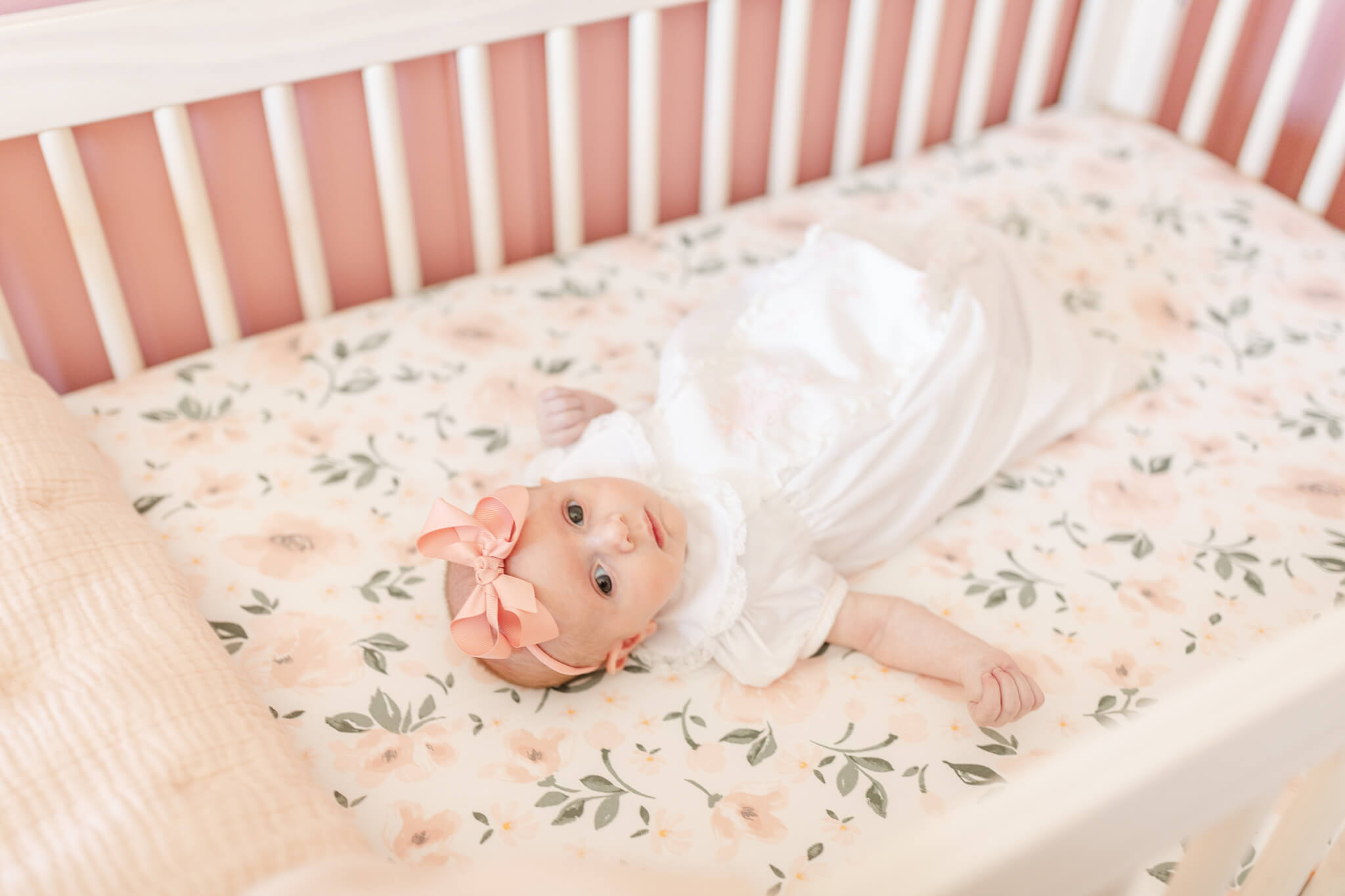 A newborn baby girl in a white dress lays in a flower print crib looking out