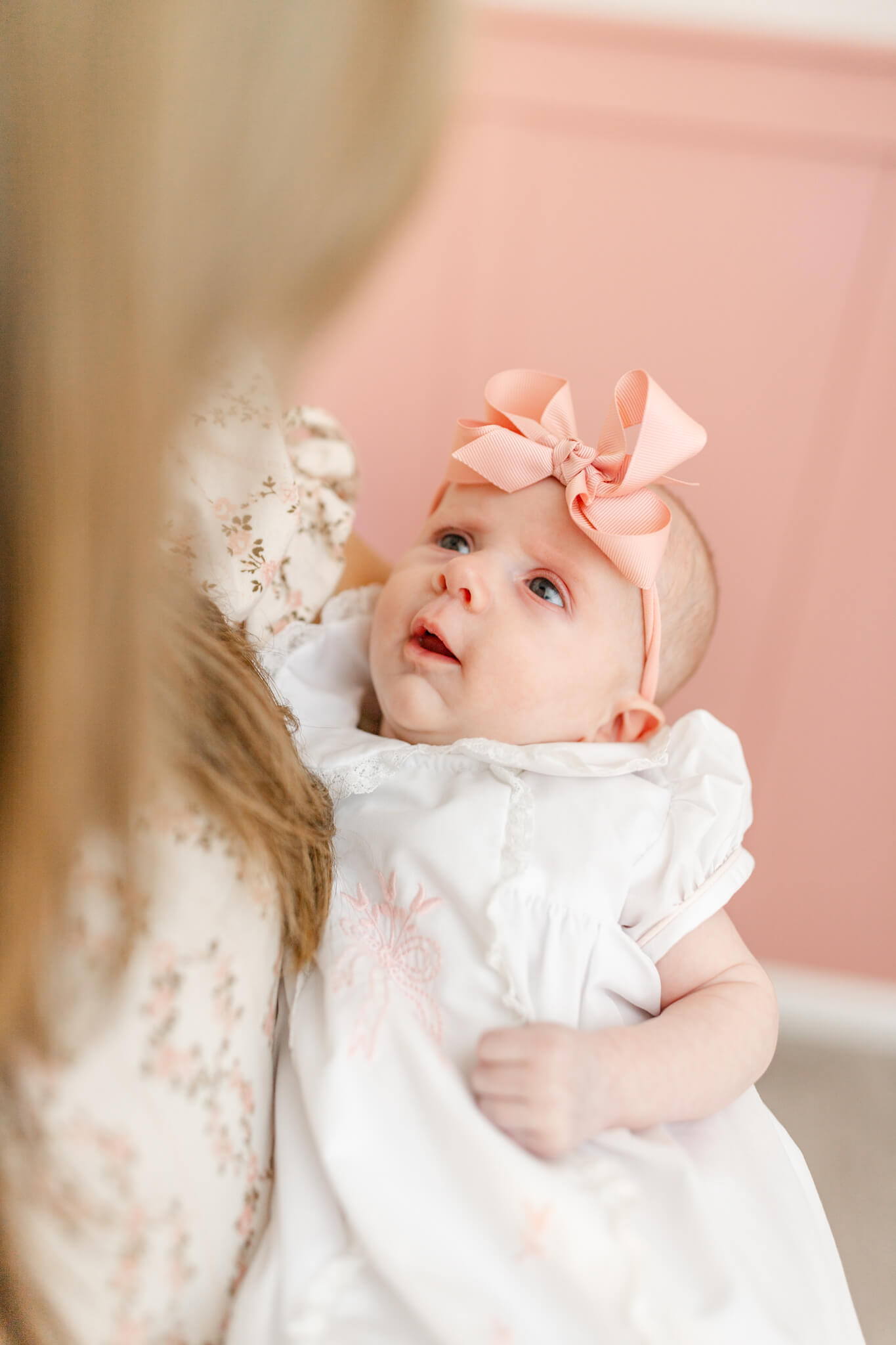 A newborn baby girl in a white dress and peach bow looks up to momma while laying in her arms after visiting baby stores in Huntsville, AL