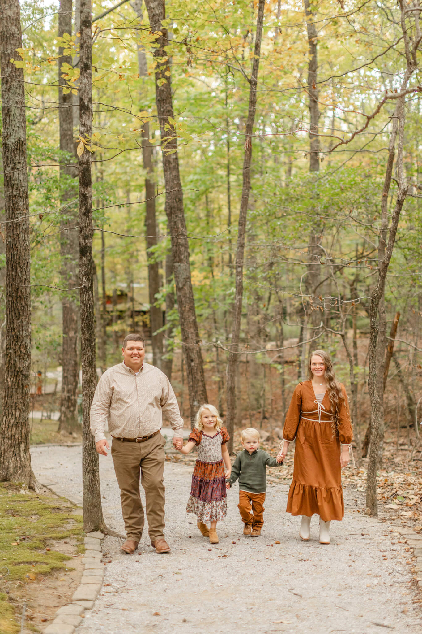 Smiling toddler brother and sister in rust and green hold hands with each other and mom and dad in a park trail after visiting boutiques in Huntsville, AL