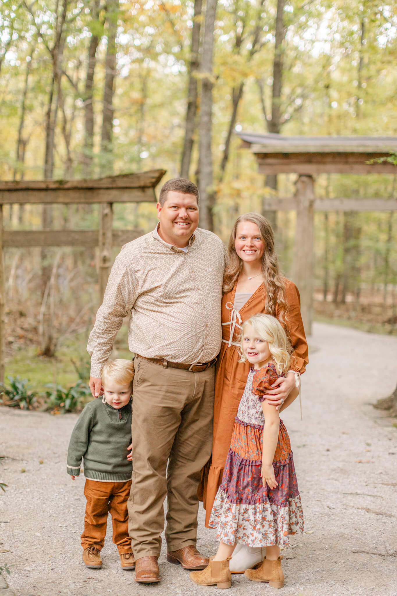 Happy mom and dad stand in a park trail smiling with their toddler son and daughter at their sides after visiting boutiques in Huntsville, AL