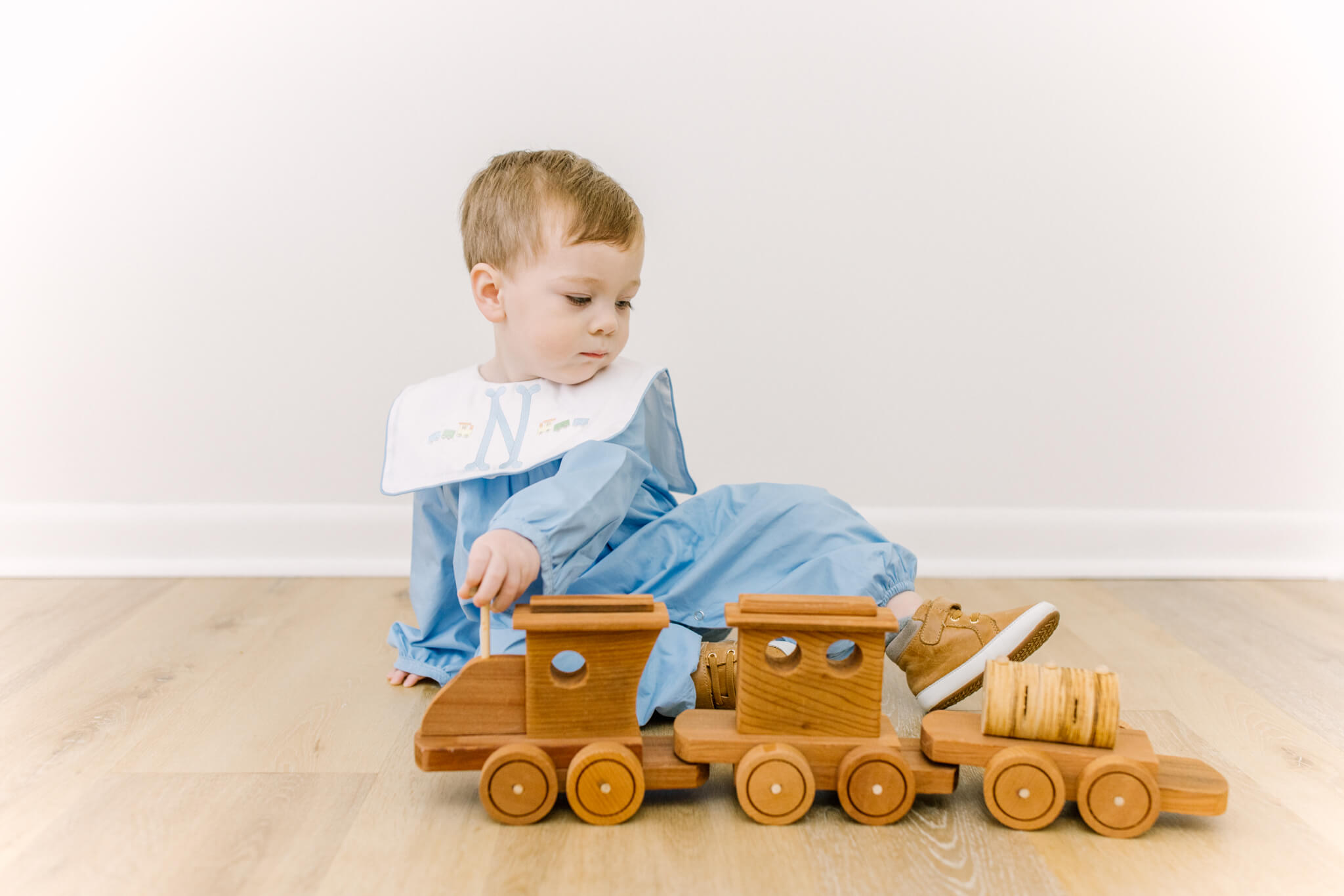 A toddler boy plays with a wooden trail in a studio in a blue romper