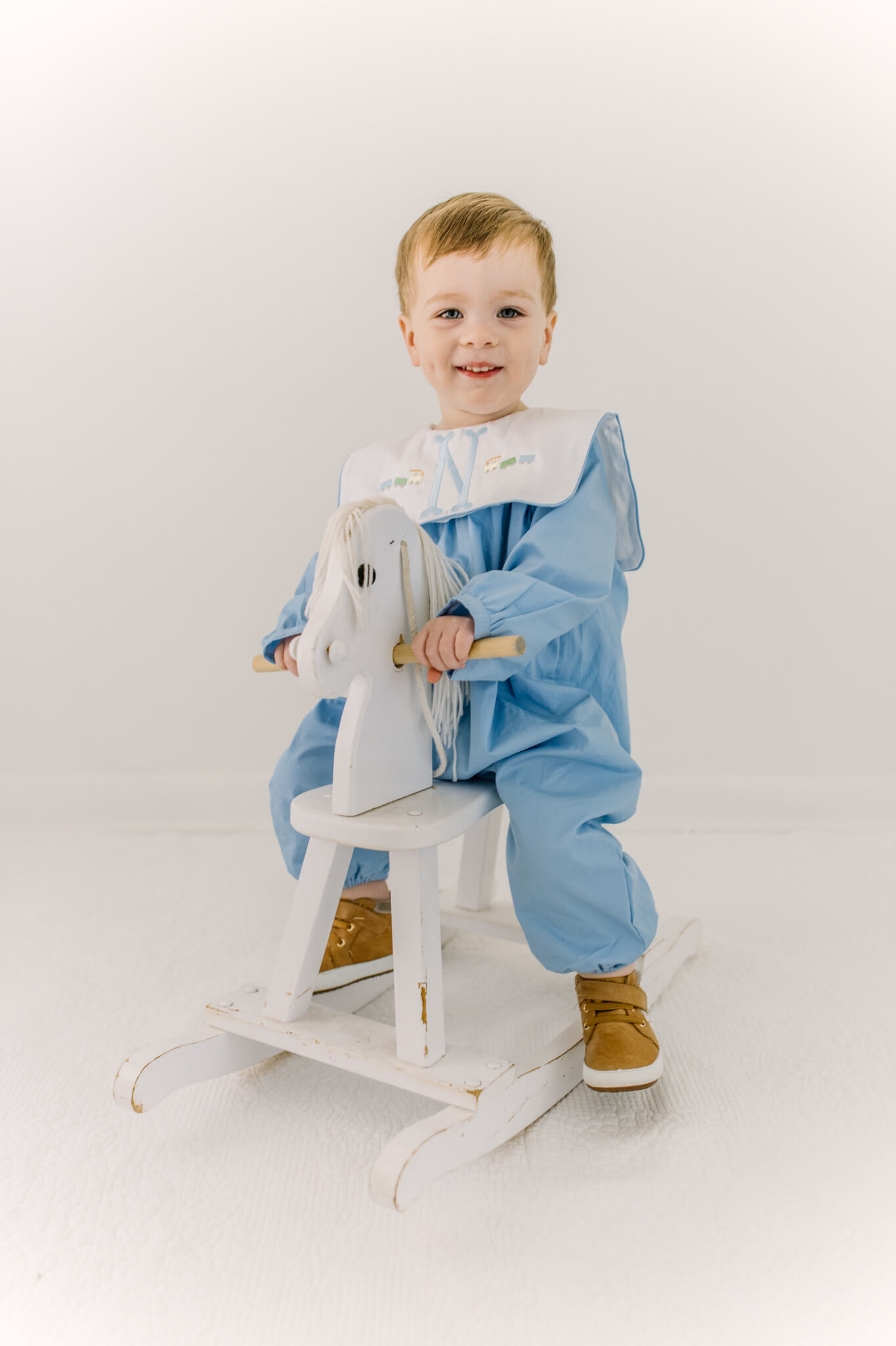A happy toddler boy rides a wooden rocking horse in a studio in a blue romper with bib before visiting indoor playgrounds in huntsville, AL