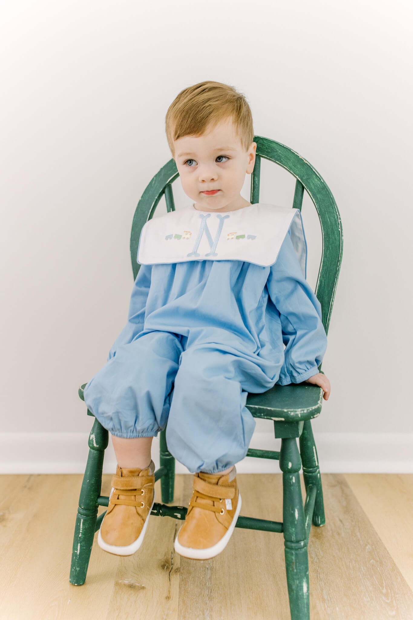 A toddler boy sits in a wooden chair in a blue romper and brown shoes before visiting indoor playgrounds in huntsville, AL