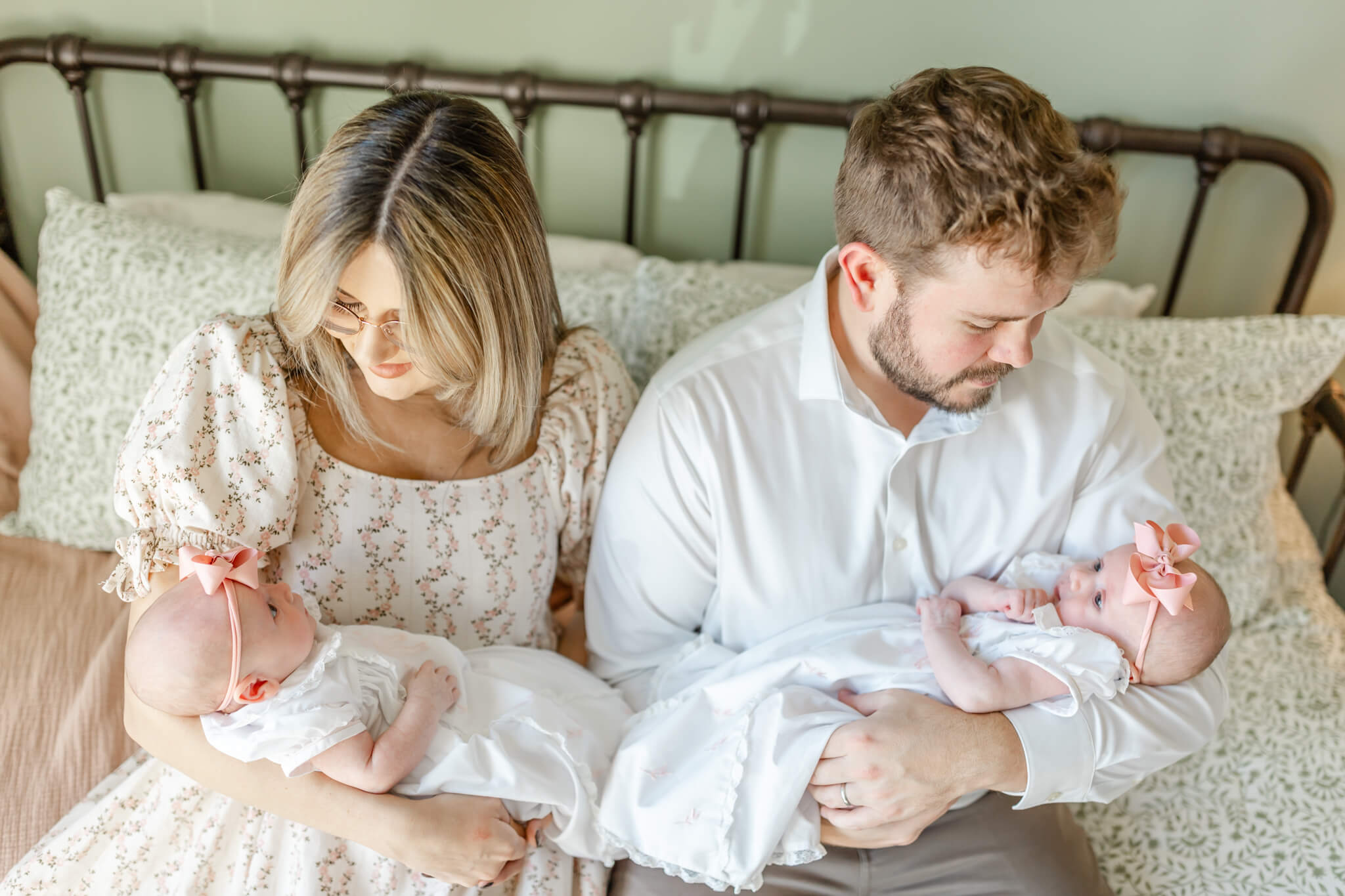 Happy new mom and dad sit on a bed cradling their newborn twin daughters in white dresses in their laps