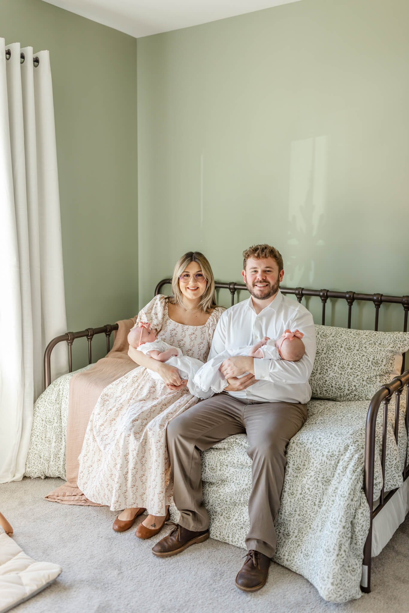 Happy new parents of twins sit on a daybed with their newborns in their arms in white dresses after meeting nannies in Huntsville, AL