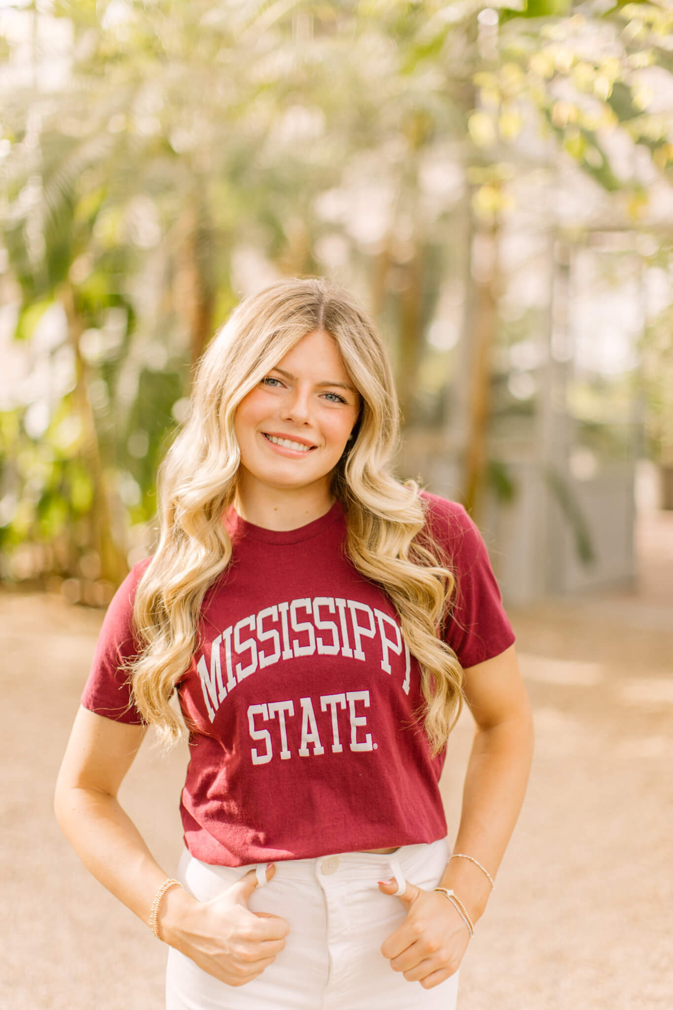 A smiling graduate stands in a garden in a maroon college shirt with fingers in belt loops
