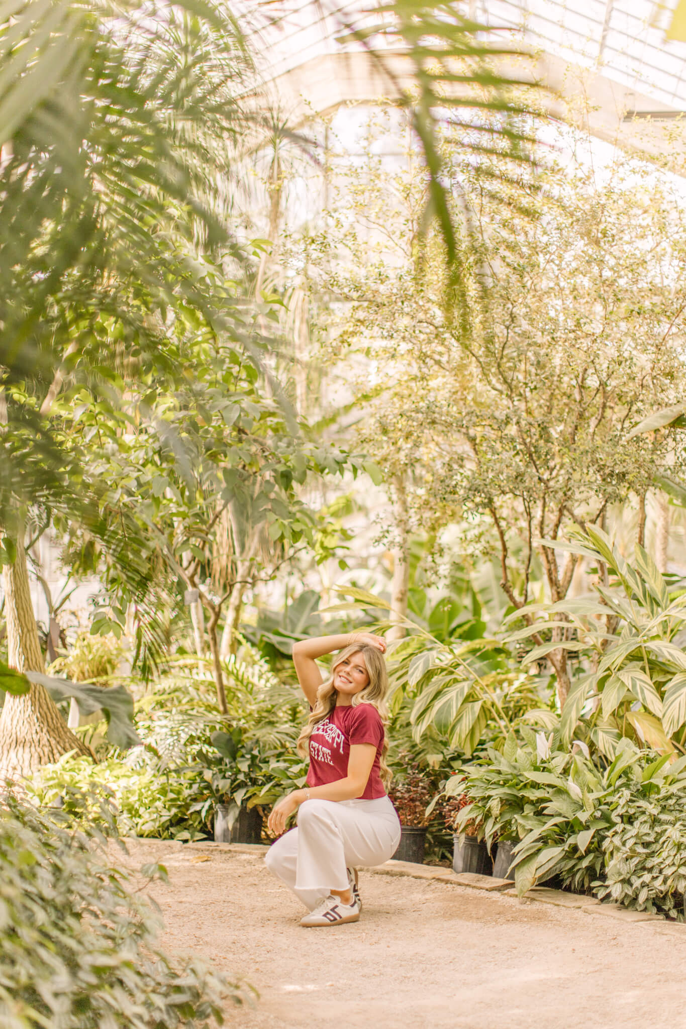 A high school senior laughs while kneeling in a greenhouse in a red shirt and white pants after visiting hair salons in Huntsville, AL