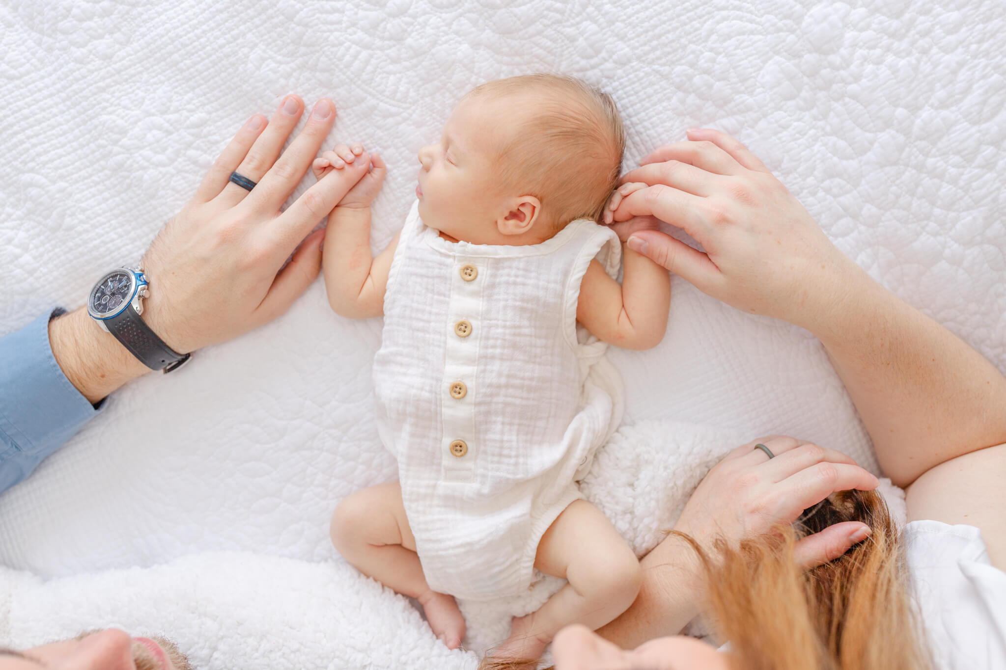 A look down at a sleeping newborn in a white onesie on a white bed holding mom and dad's fingers