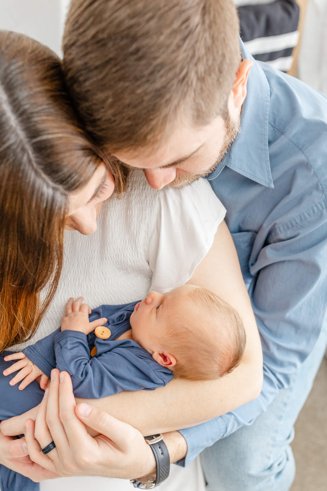 A newborn baby lays in mom and dad's arms as they snuggle and look down to him in a blue onesie thanks to health first chiropractic in huntsville, AL