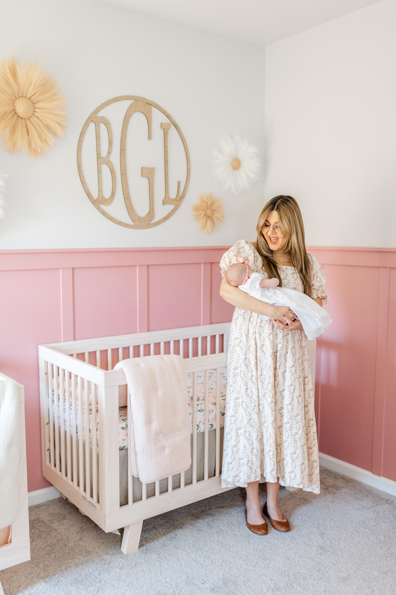 A happy mom laughs while holding her newborn in a nursery by crib with pink and white walls