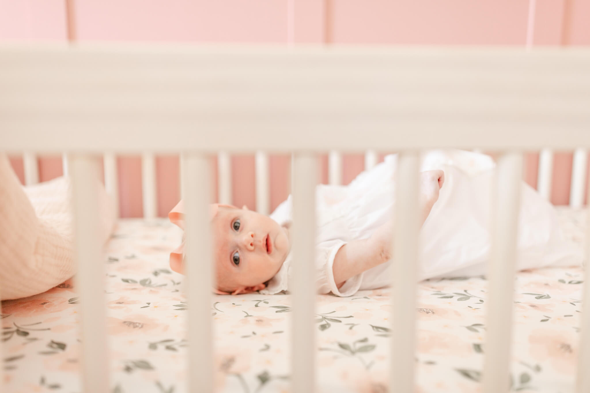 A newborn baby lays in a crib in a white dress and pink bow with eyes open after meeting lactation consultants in Huntsville, AL