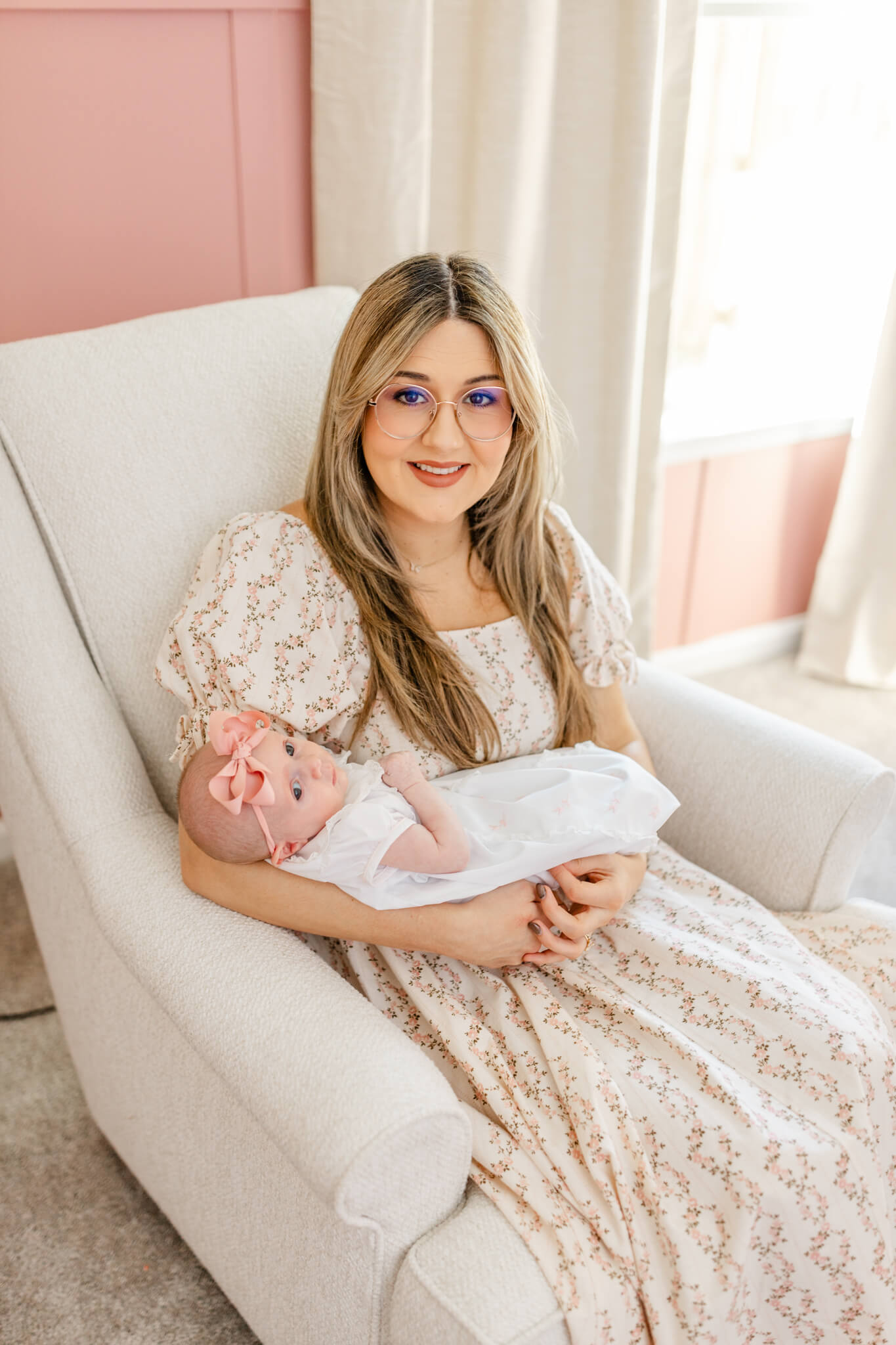 A happy new mom sits in a nursing chair holding her newborn in a white dress after meeting lactation consultants in Huntsville, AL