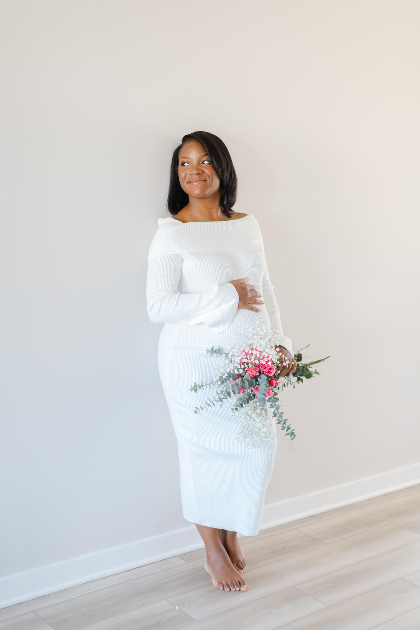 A pregnant woman leans against a wall holding a pink rose bouquet in a white maternity dress
