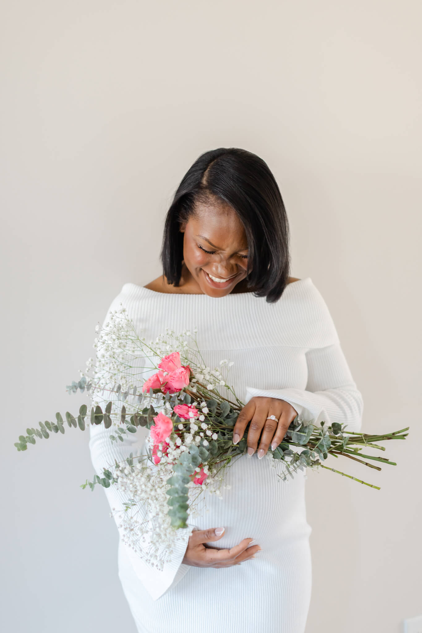 A laughing mother to be stands in a studio in a white dress holding pink roses after meeting midwives in Huntsville, AL