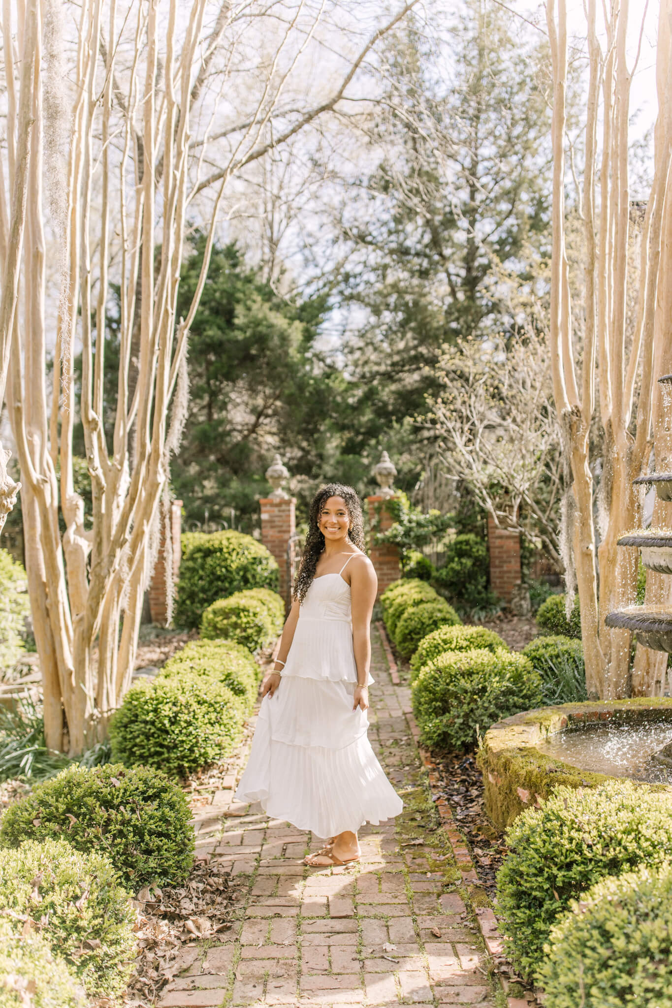 A smiling high school senior in a white dress twirls in a garden path
