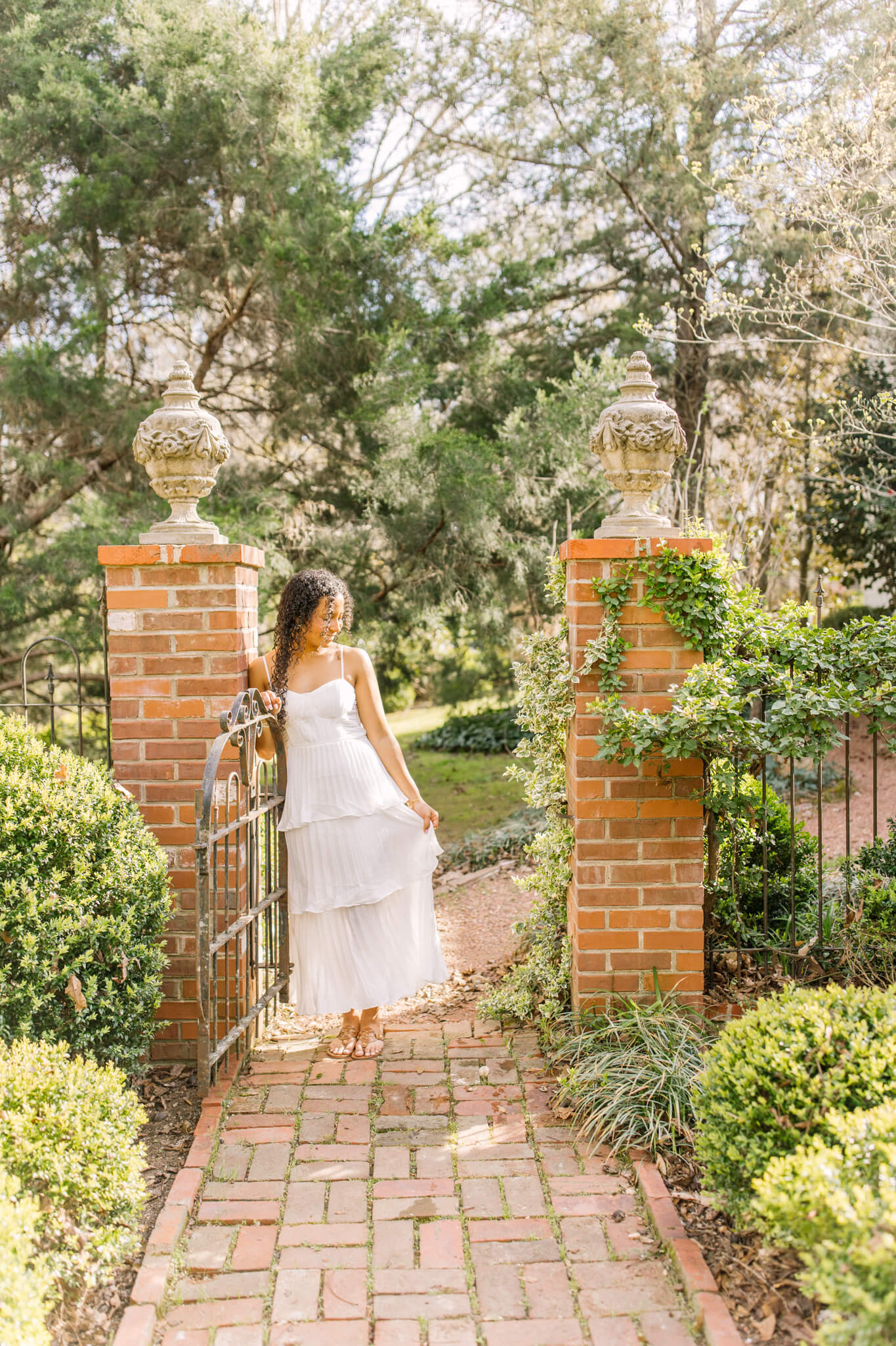 A high school senior in a white dress smiles while leaning in a garden entrance after shopping for prom dresses in huntsville al