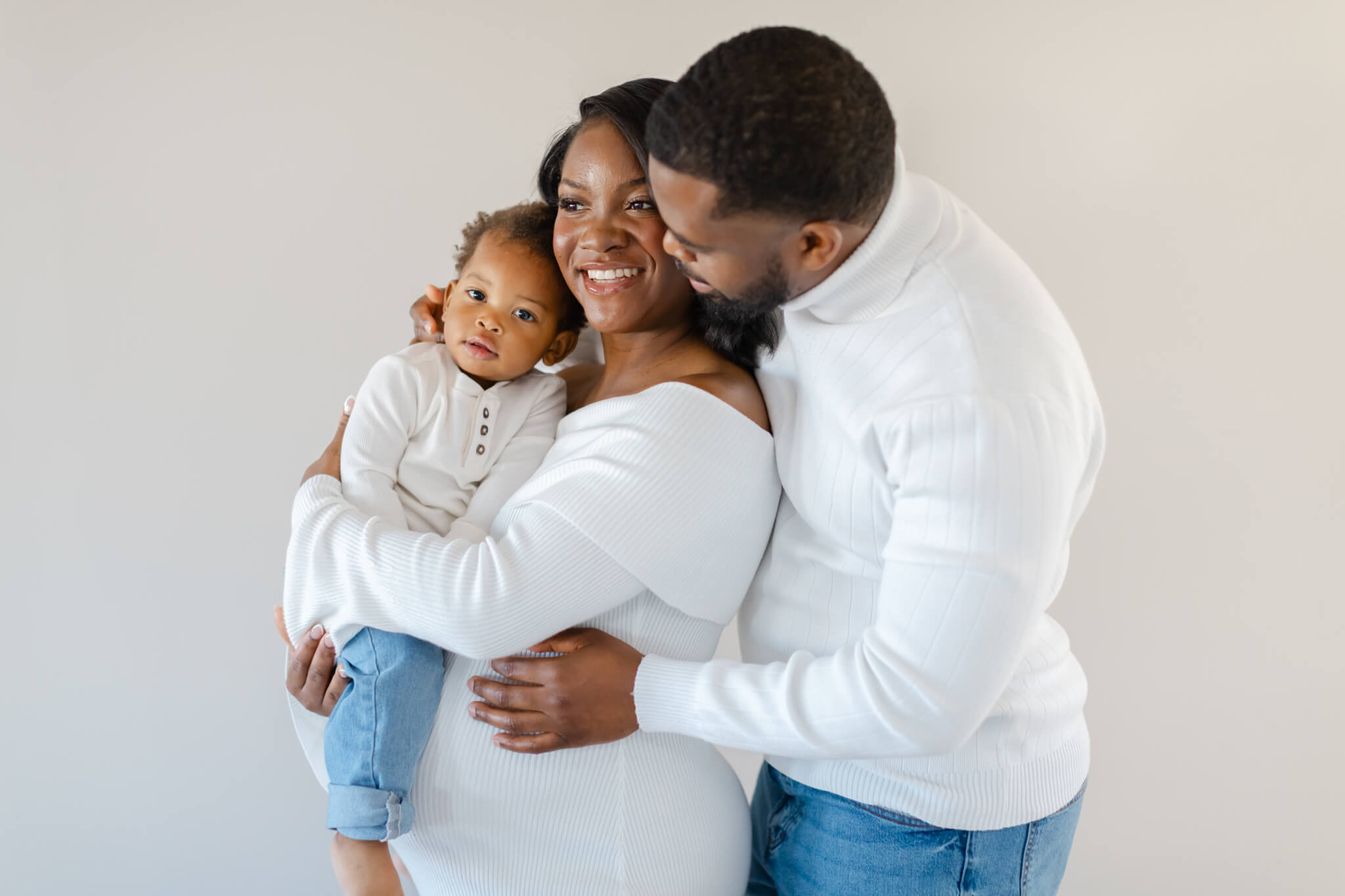 A toddler boy sits on mom's pregnant belly as dad snuggles her from behind in white outfits