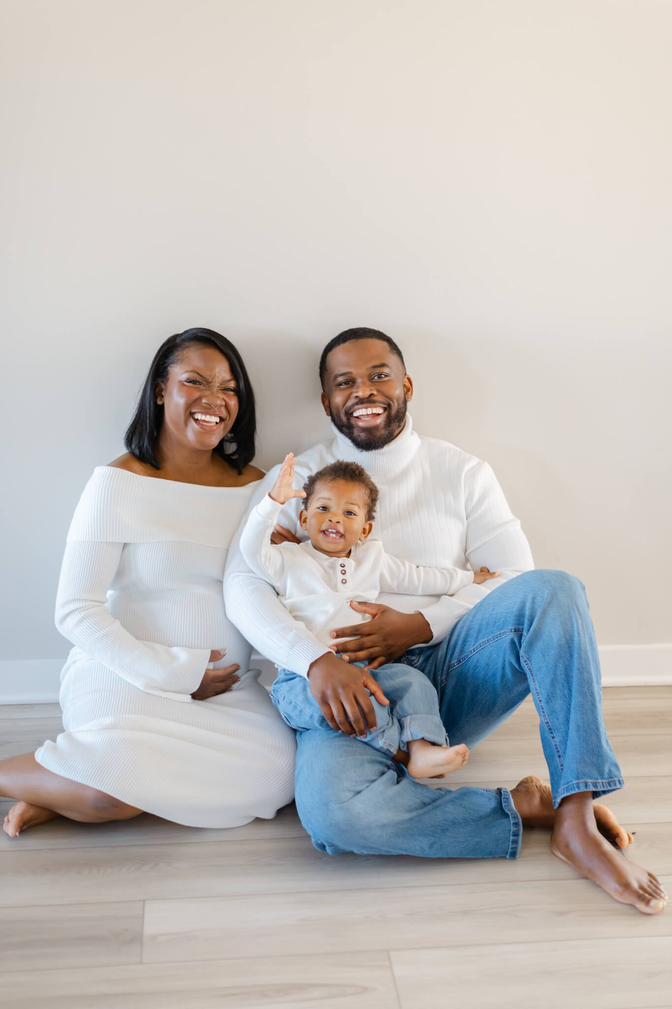A smiling mom and dad in white and denim sit on the floor witht heir toddler son in their lap after meeting doulas in Huntsville, AL