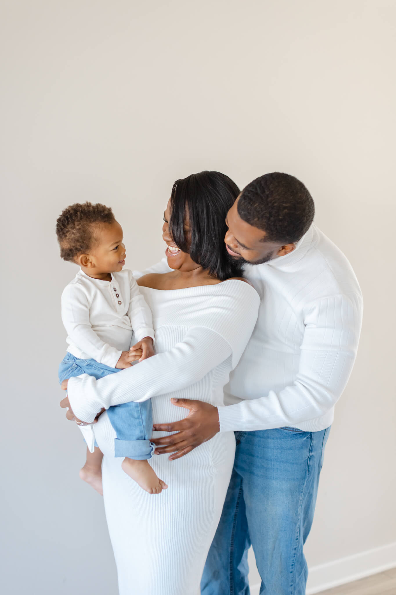 Happy mom and dad laugh with their toddler son on mom's pregnant belly while standing in white and denim after meeting doulas in Huntsville, AL