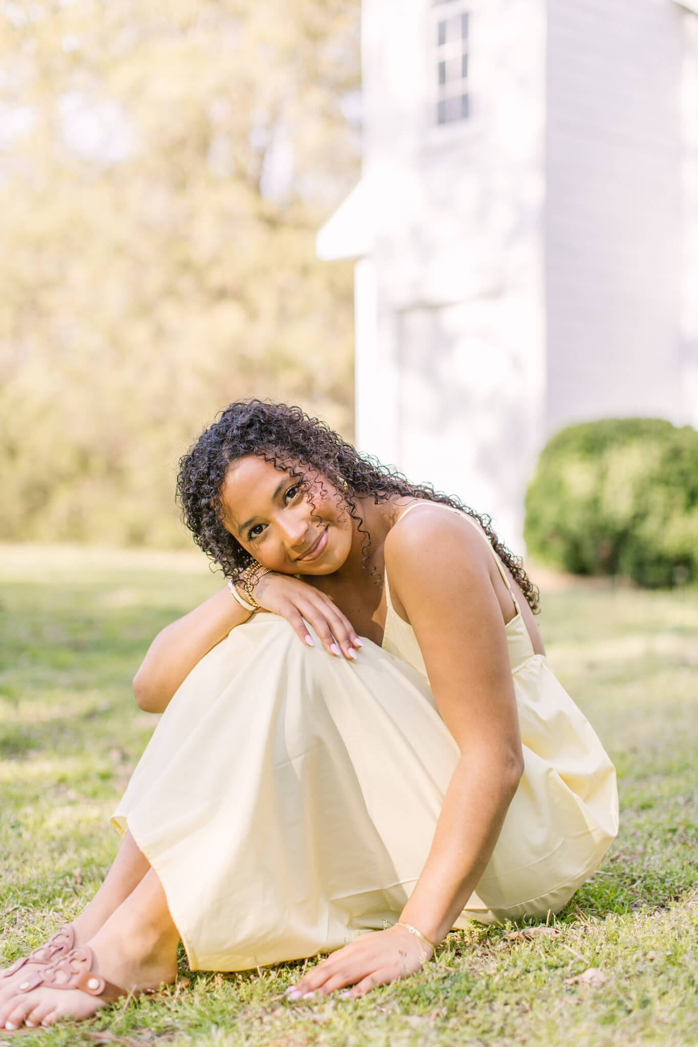A happy high school senior sits in a lwn wearing a yellow dress leaning on her knees with smiles