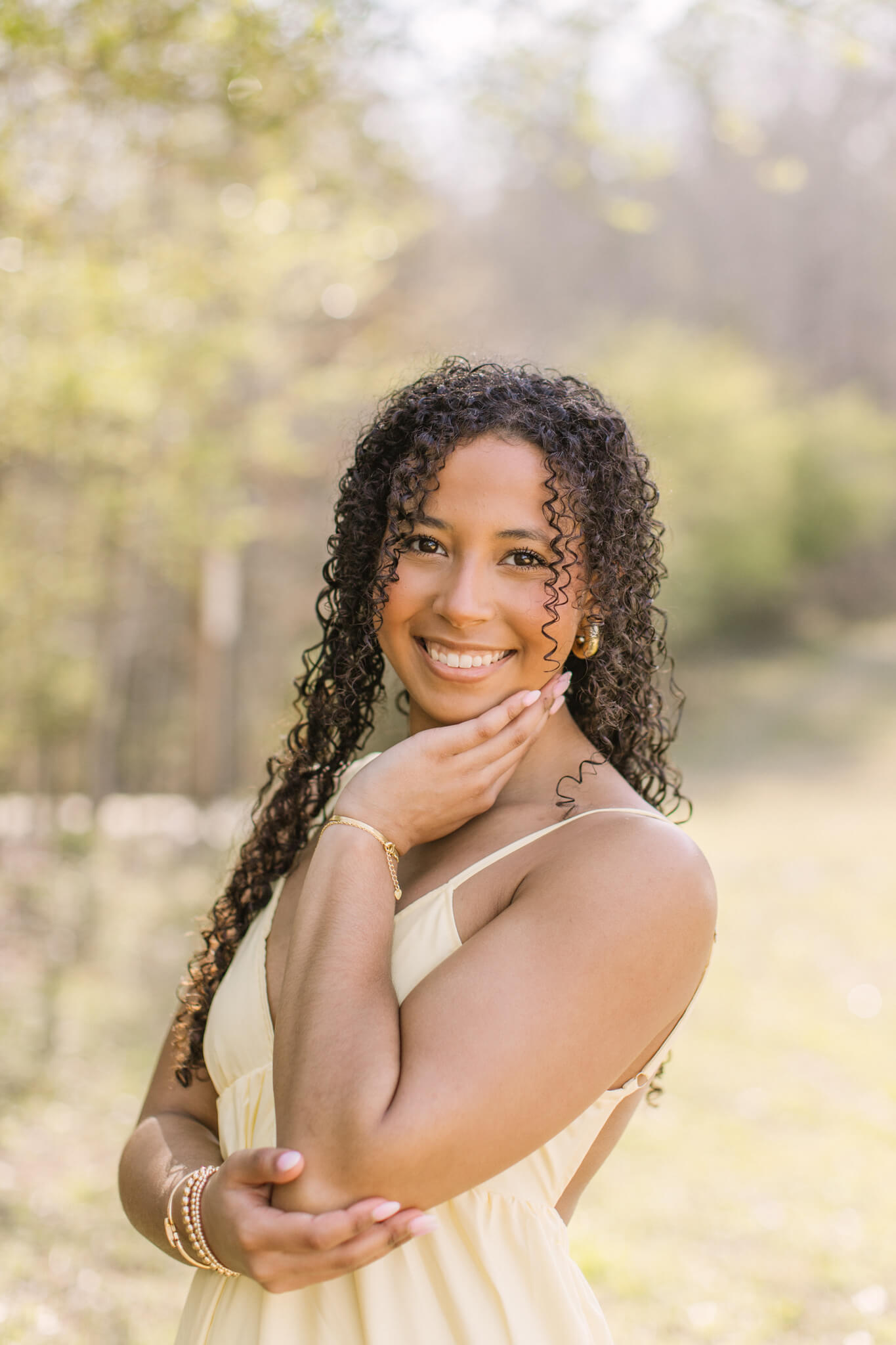 A smiling high school senior rests her chin in her hand while standing in a lawn at sunset in a yellow dress after finding great nail salons in Huntsville, AL