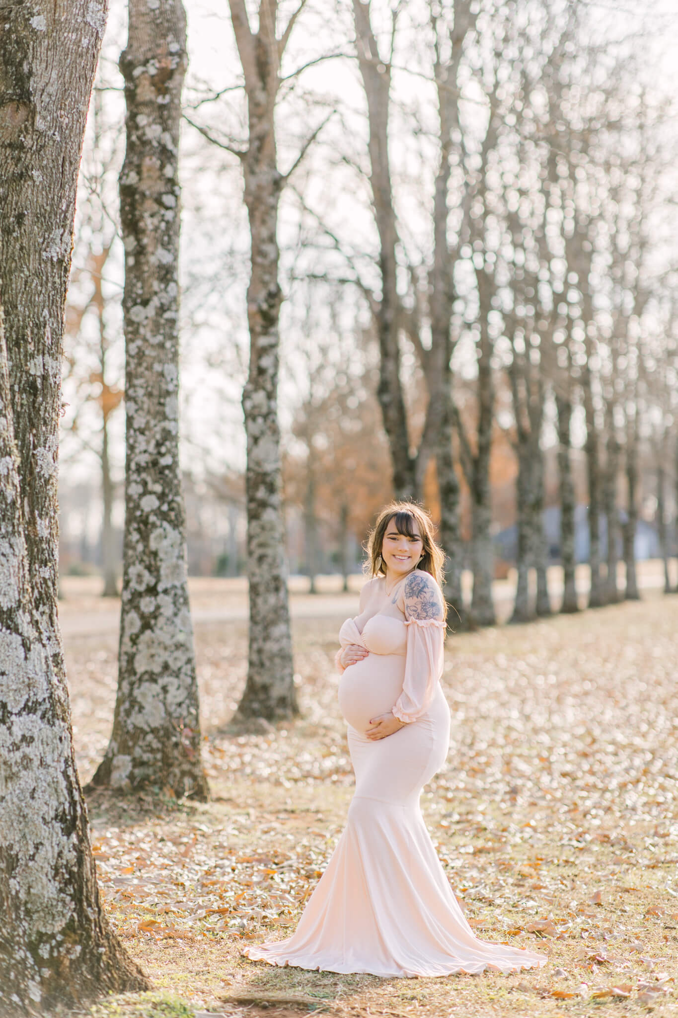A smiling expecting woman in a pink maternity gown stands in a row of trees in fall at sunset smiling