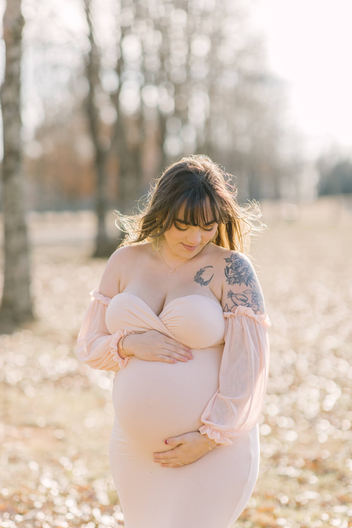 A smiling pregnant woman in a pink body con maternity gown looks down to her hands on her bump in a field at sunset after some pelvic floor therapy in Huntsville, AL