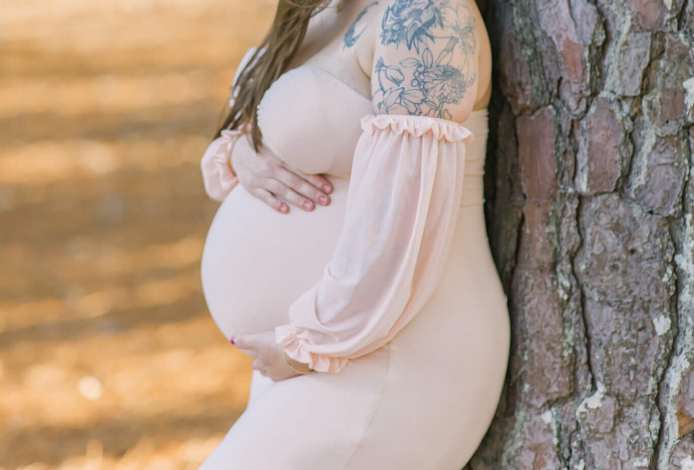 A mother to be in a pink maternity gown leans against a pine tree gazing down her shoulder after some pelvic floor therapy in Huntsville, AL
