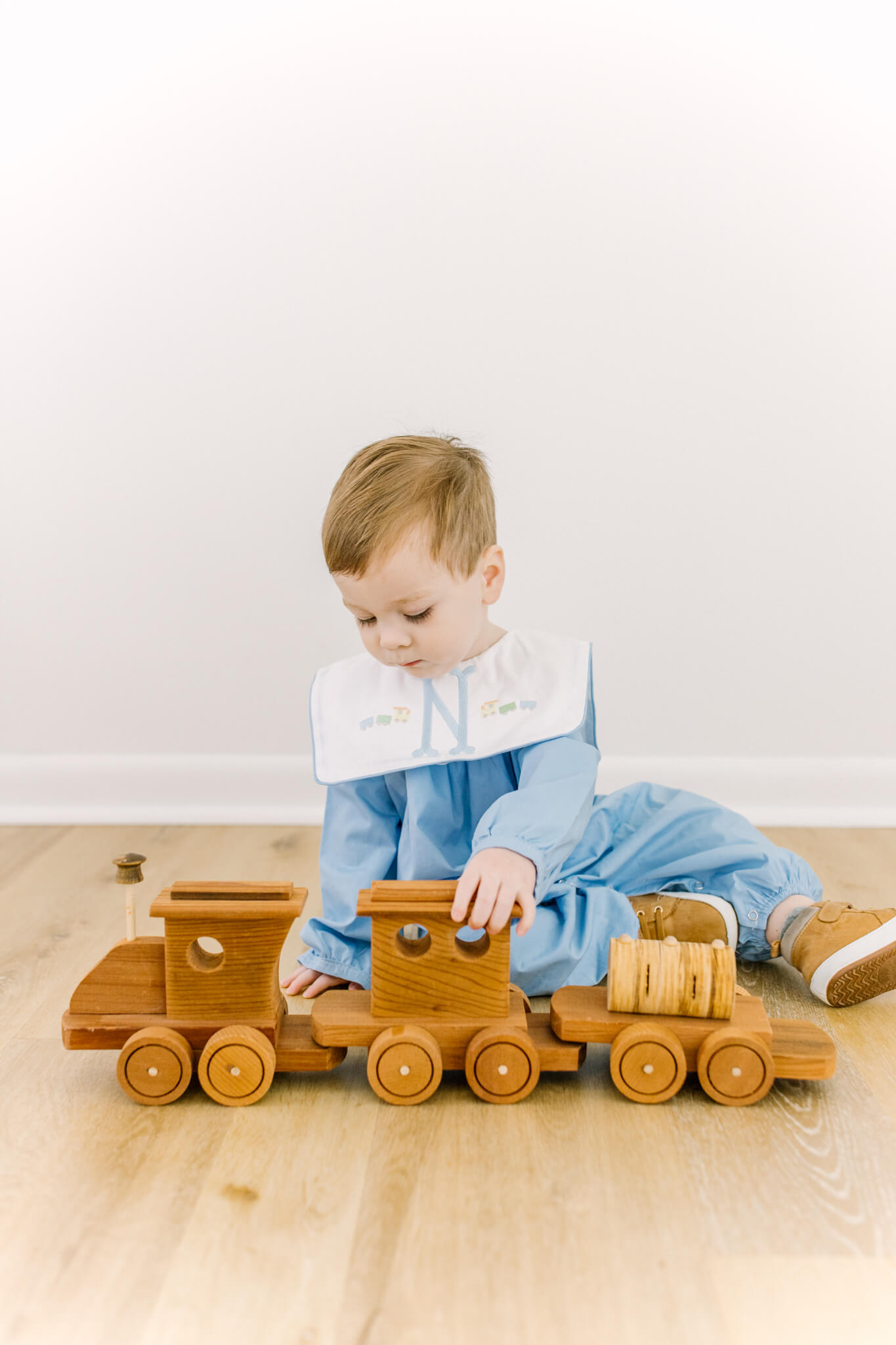 A toddler boy plays with a wooden train on the floor while wearing a blue heirloom onesie after visiting toy stores in Huntsville, AL