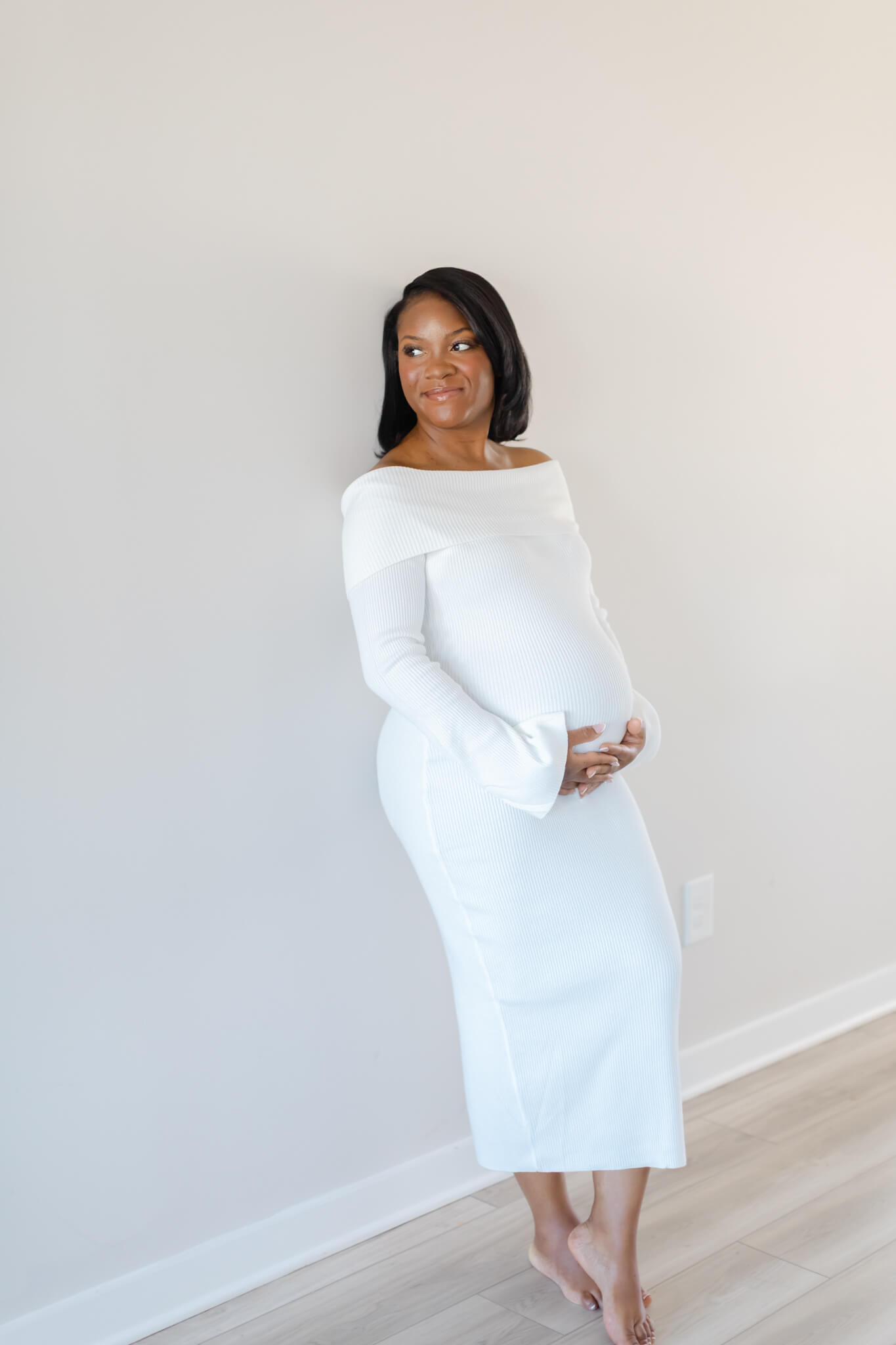 A smiling pregnant woman leans against a white wall in a hallway holding her bump in a white maternity gown