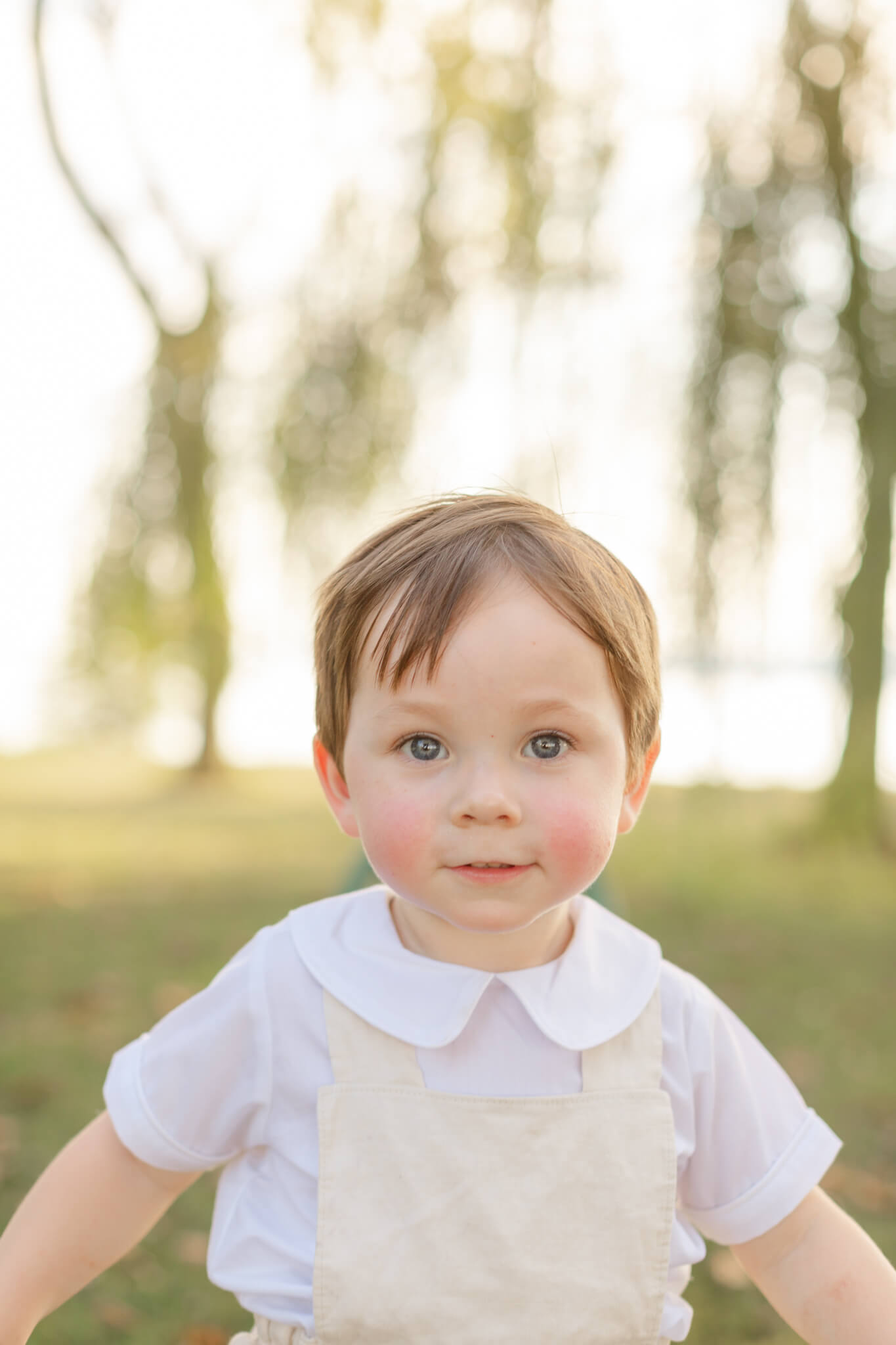A toddler smiles while in yellow overalls in a park with rosy cheeks