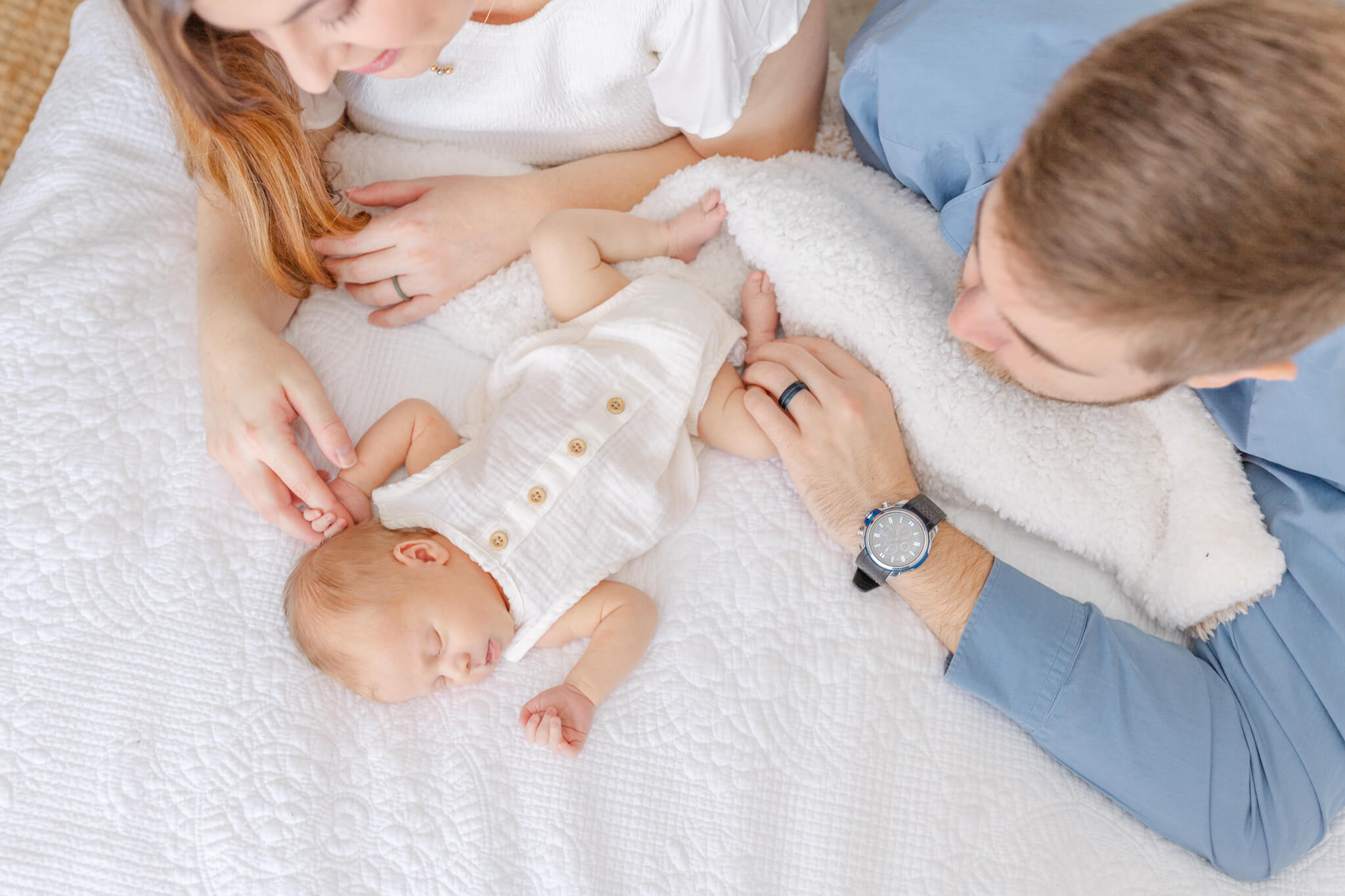 A newborn baby sleeps on a bed in a white onesie as mom and dad admire him