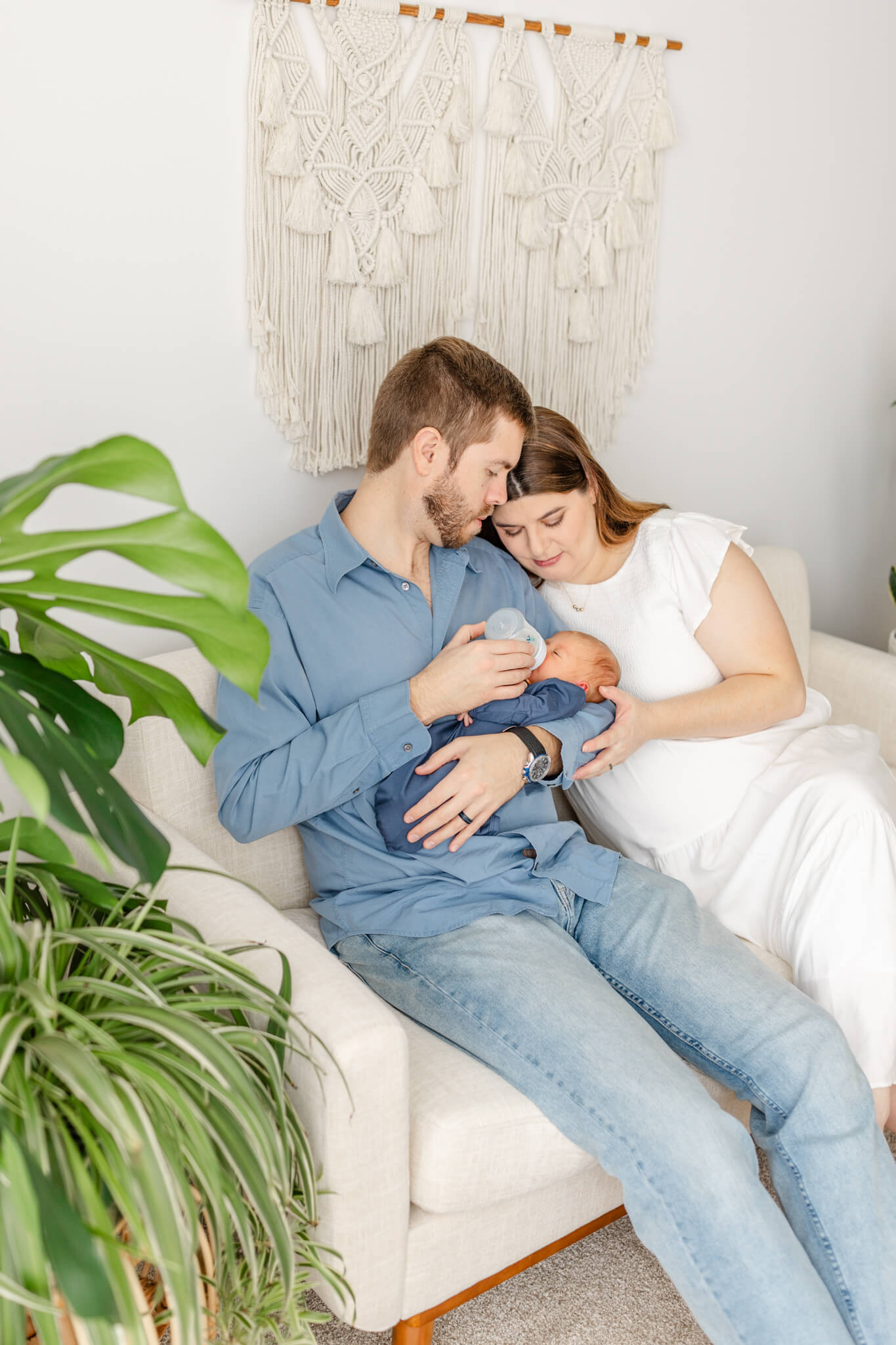 NEw mom and dad sit on a couch feeding their newborn in a blue onesie thanks to postpartum doulas in Huntsville, AL