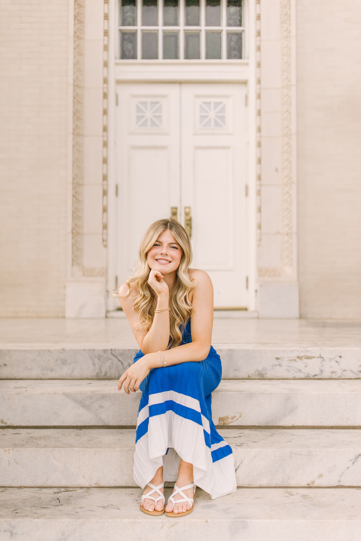 A happy high school senior sits on marble steps in a blue dress leaning on her knee