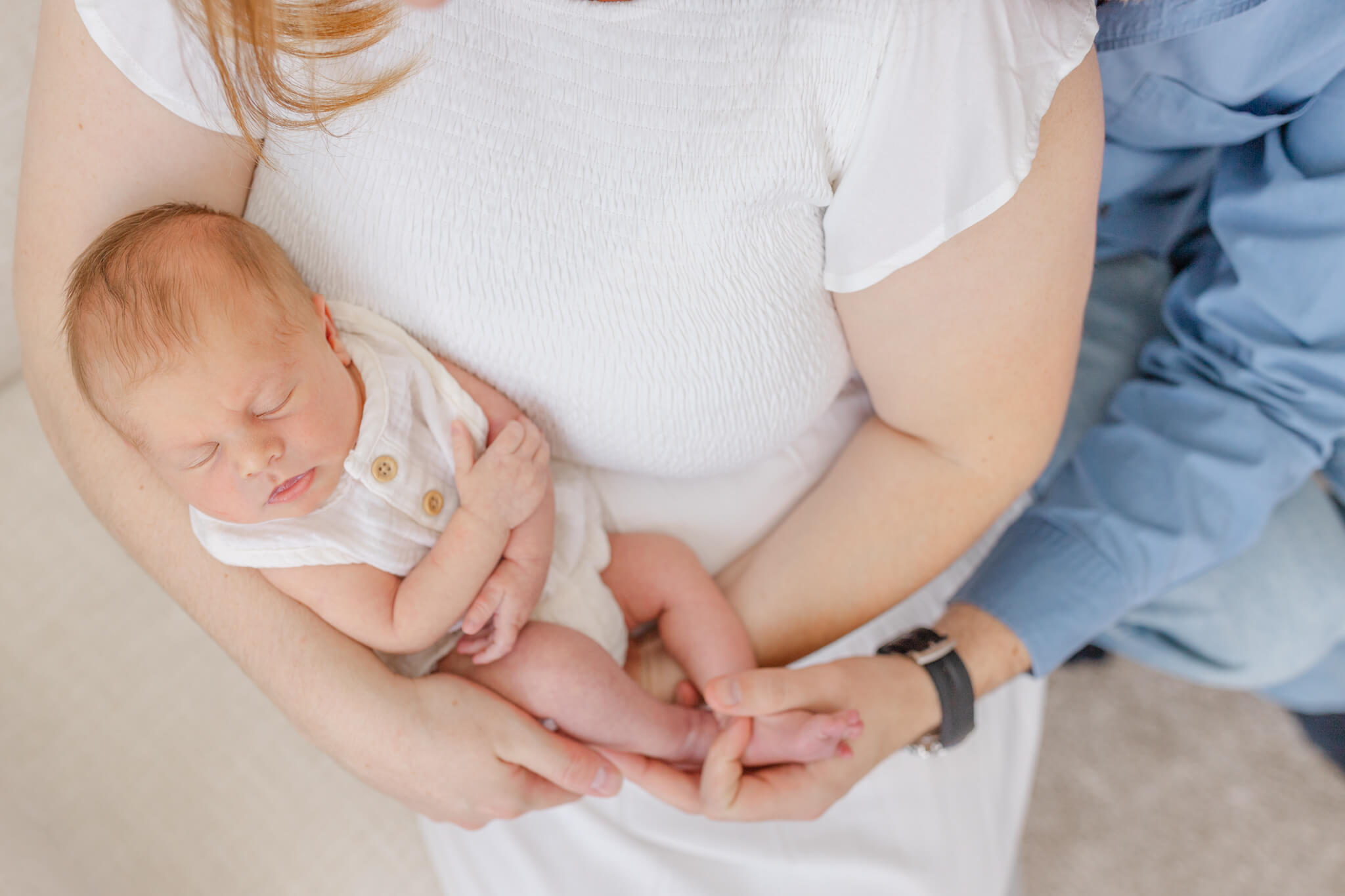 A newborn baby sleeps in mom's arms in a white onesie as dad holds the feet