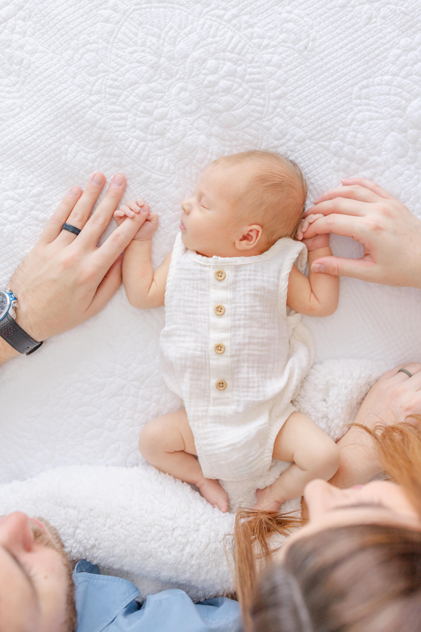 A newborn baby sleeps in a white onesie while holding mom and dad's fingers thanks to sleep coaching in Huntsville, AL