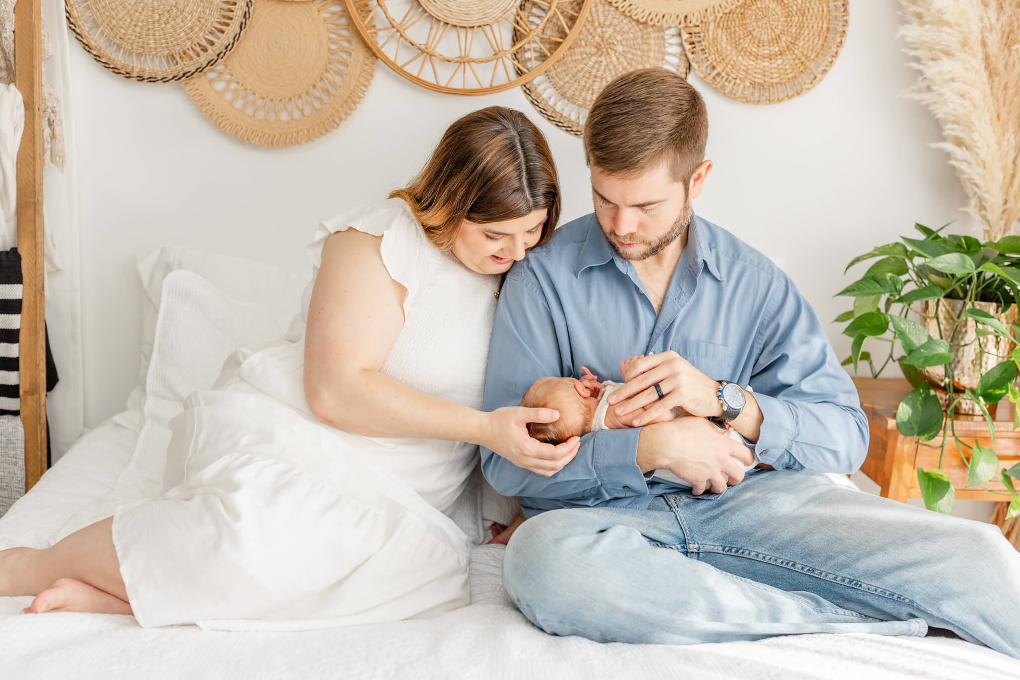 New mom and dad in blue and white sit on a bed cradling their newborn baby in dad's lap after some sleep coaching in Huntsville, AL