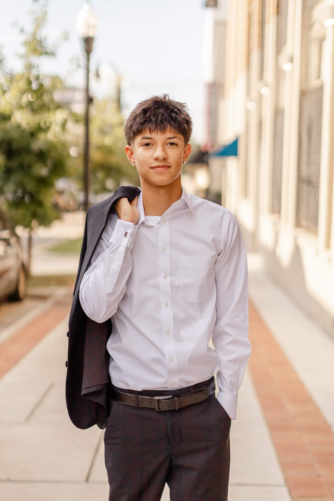 A teenage boy in a white button down and slacks holds his suit jacket over his shoulder while walking on a downtown sidewalk