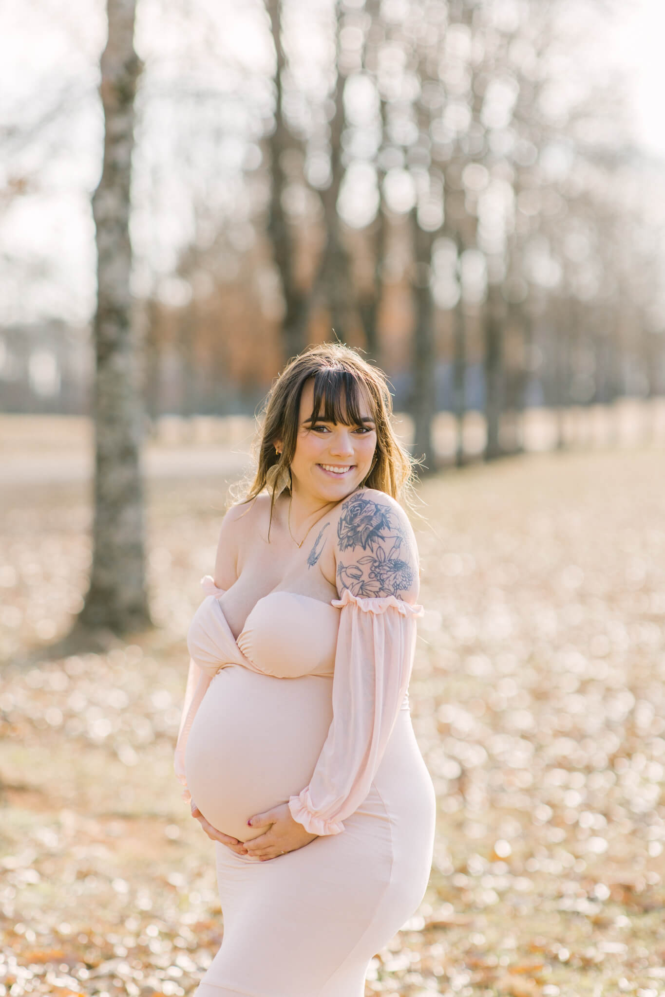 A happy new mom in a pink maternity gown smiles while walking in a park at sunset