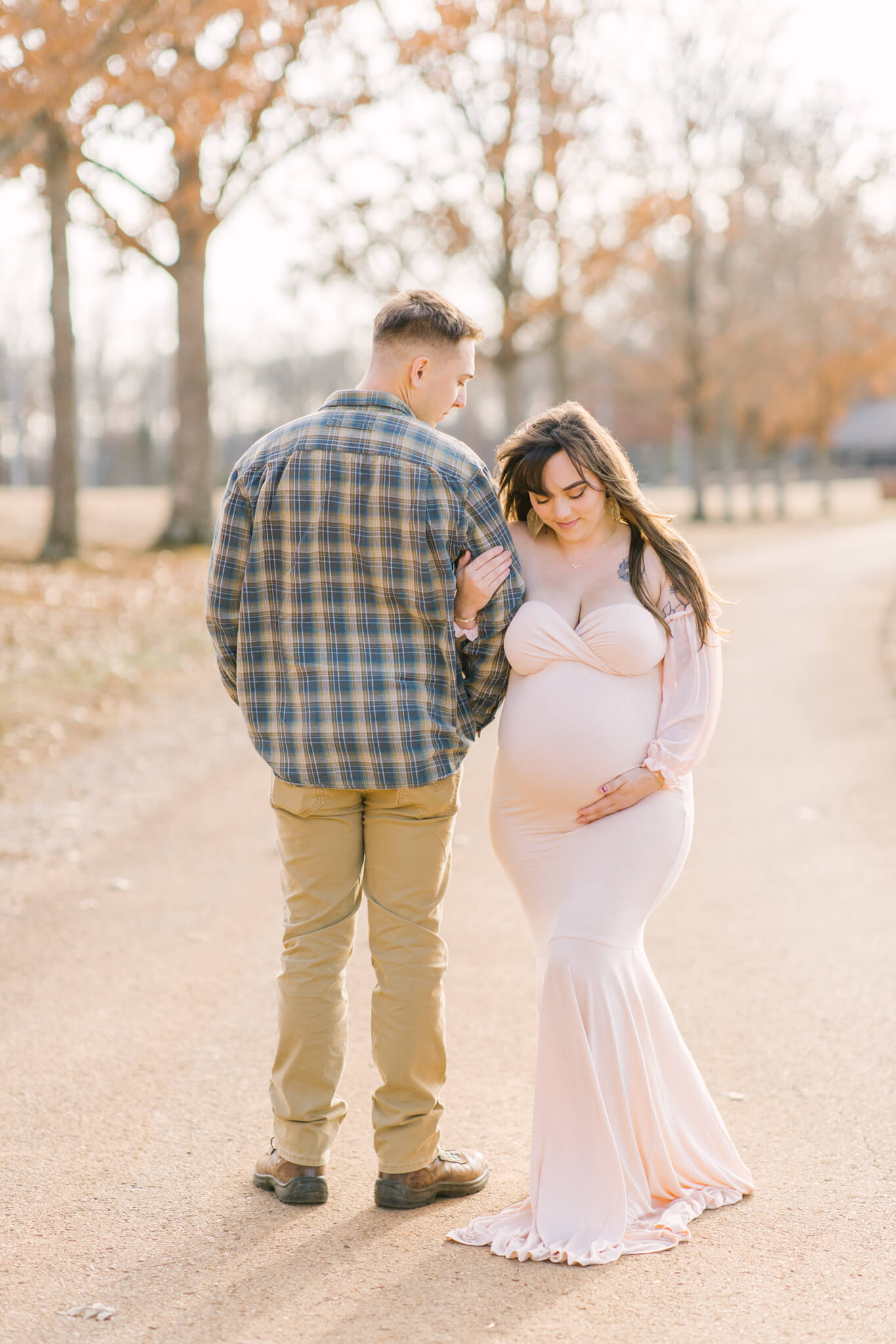 A happy pregnant couple stand in a park sidewalk at sunset as she smiles down to her bump in a pink maternity gown after visiting momtastic fitness