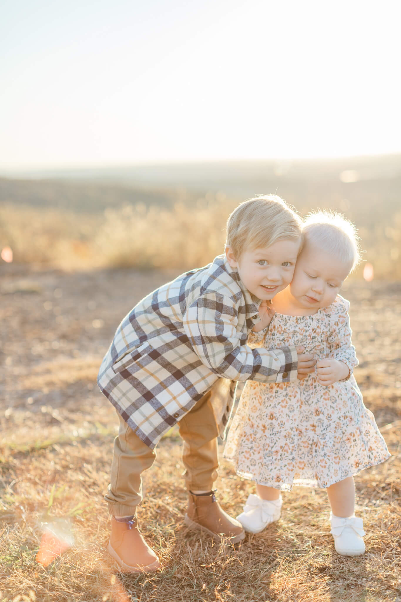 A happy toddler boy in plaid hugs his baby sister as they explore a park at sunset after visiting pediatric dentists in Huntsville, AL