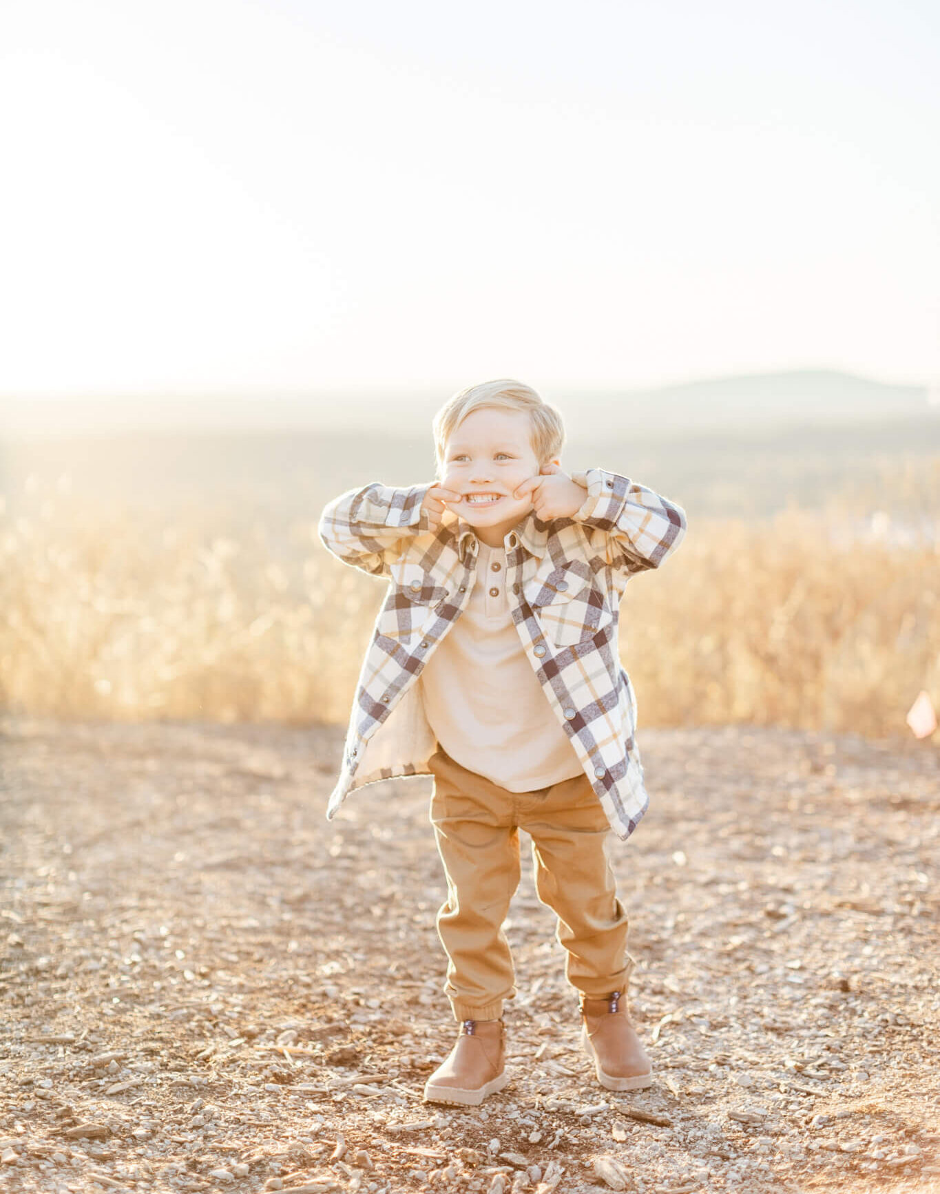 A young boy in a plaid shirt and khaki pants pulls his cheeks for a silly smile in a park at sunset thanks to pediatric dentists in Huntsville, AL